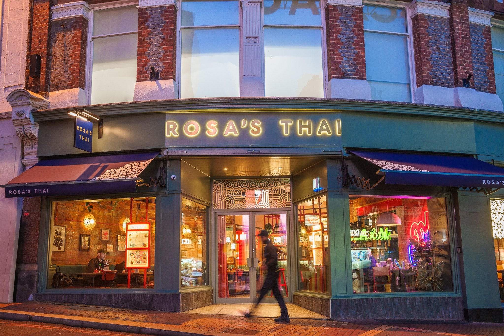 The exterior of Rosa's Thai restaurant at dusk, with large glass windows, neon signs inside, and a person walking past the entrance.