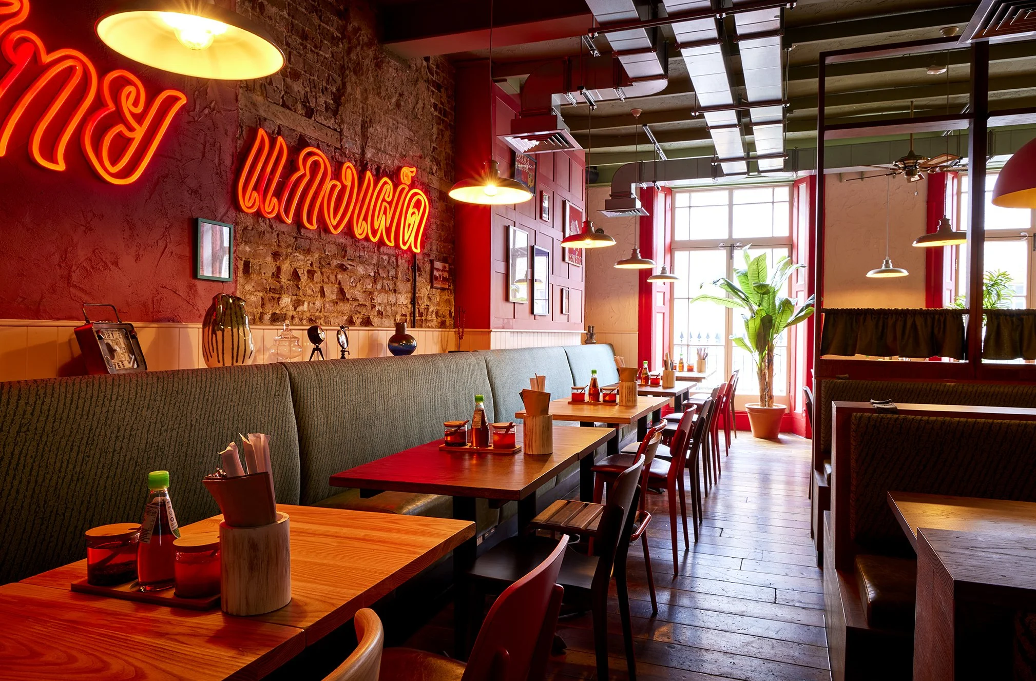 Interior of a restaurant with wooden tables, red chairs, green plant near large windows, exposed brick wall, neon signs, and hanging lights.