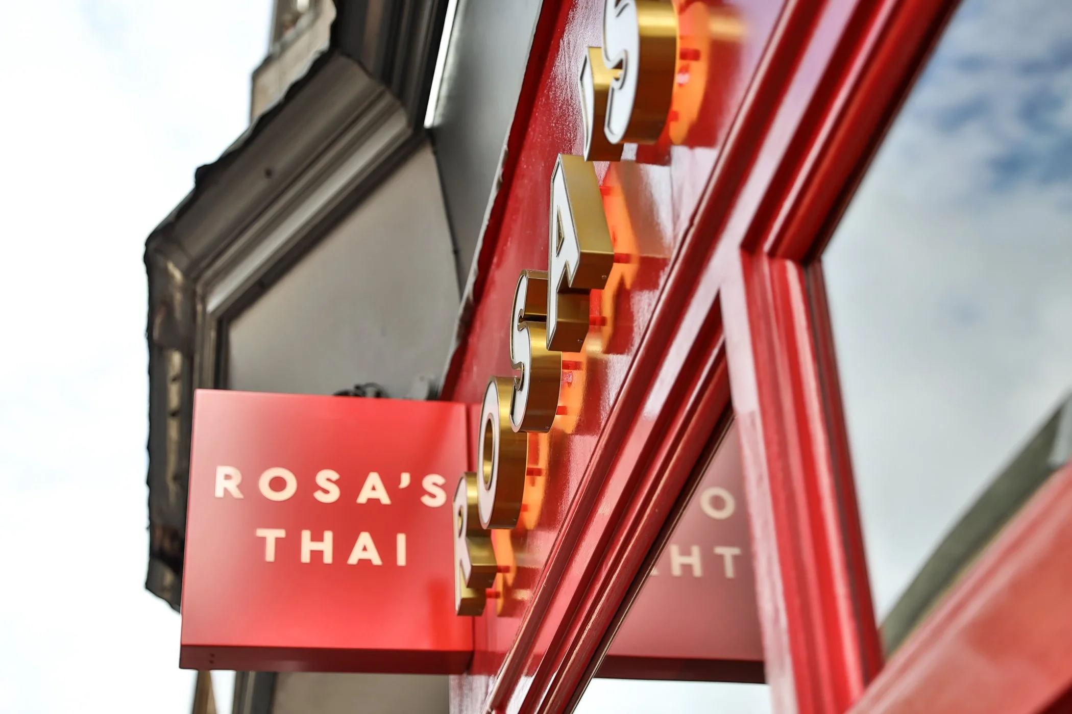 Close-up of a red and gold illuminated sign for Rosa's Thai restaurant, viewed from below against a cloudy sky.