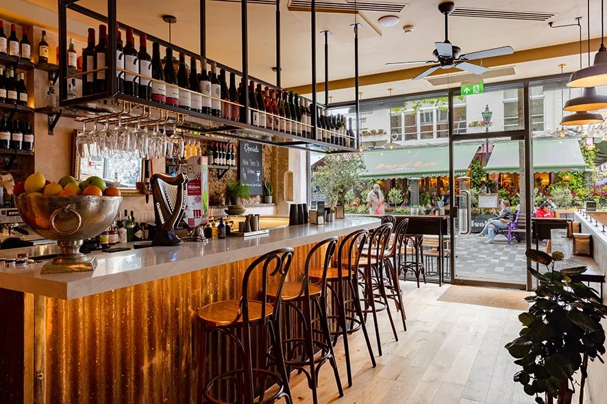 Interior of a cozy cafe with a bar counter, wine bottles on shelves, bar stools, and large windows looking out to outdoor seating area.