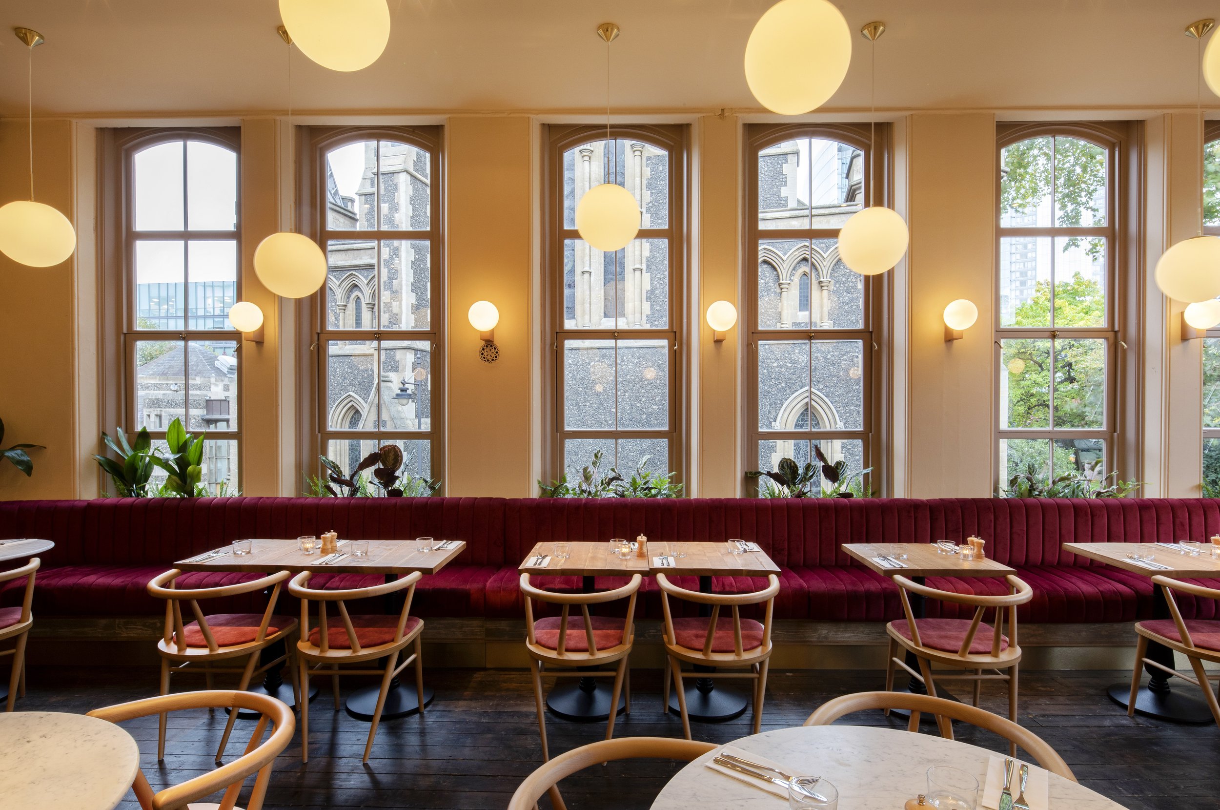 Interior of a restaurant with large arched windows, a long red velvet banquette, wooden chairs, and hanging globe pendant lights.