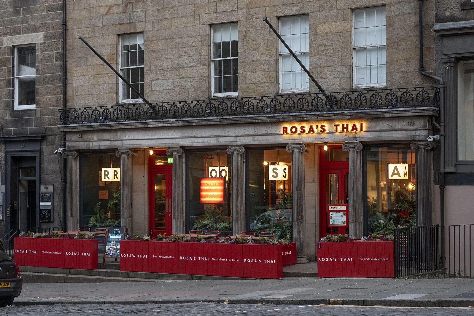Exterior of Rosa's Thai restaurant with large glass windows on a city street, red planters, and a sign overhead illuminated with the restaurant's name, showing the street and surrounding buildings.