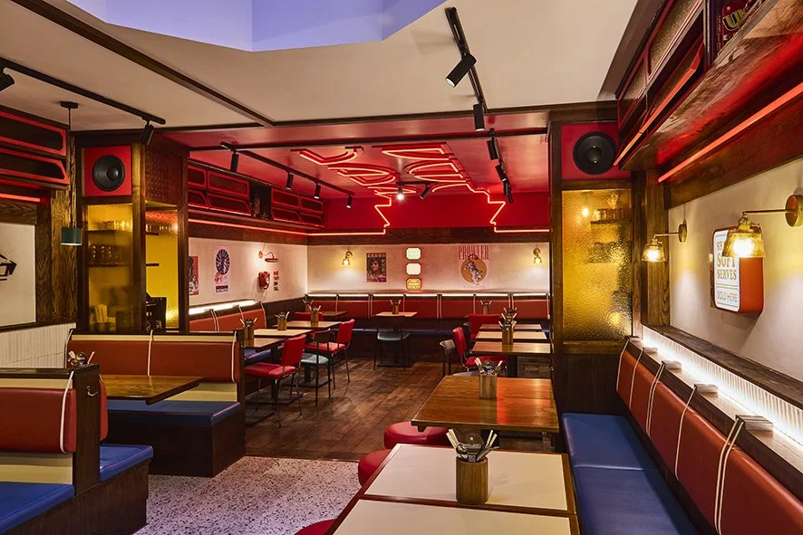 Empty restaurant with wooden tables, red and blue cushioned seating, and retro-style lighting, neon signs, and decor.