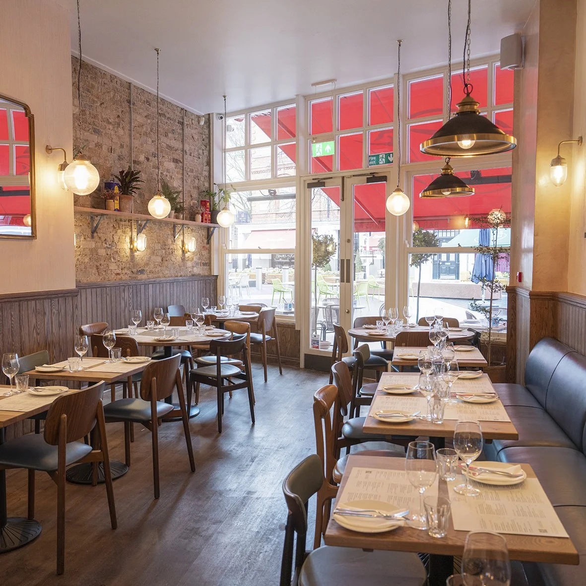 Interior of a cozy restaurant with wooden tables, chairs, and glassware, large windows with red awnings outside, exposed brick wall, warm lighting, and a modern rustic decor style.