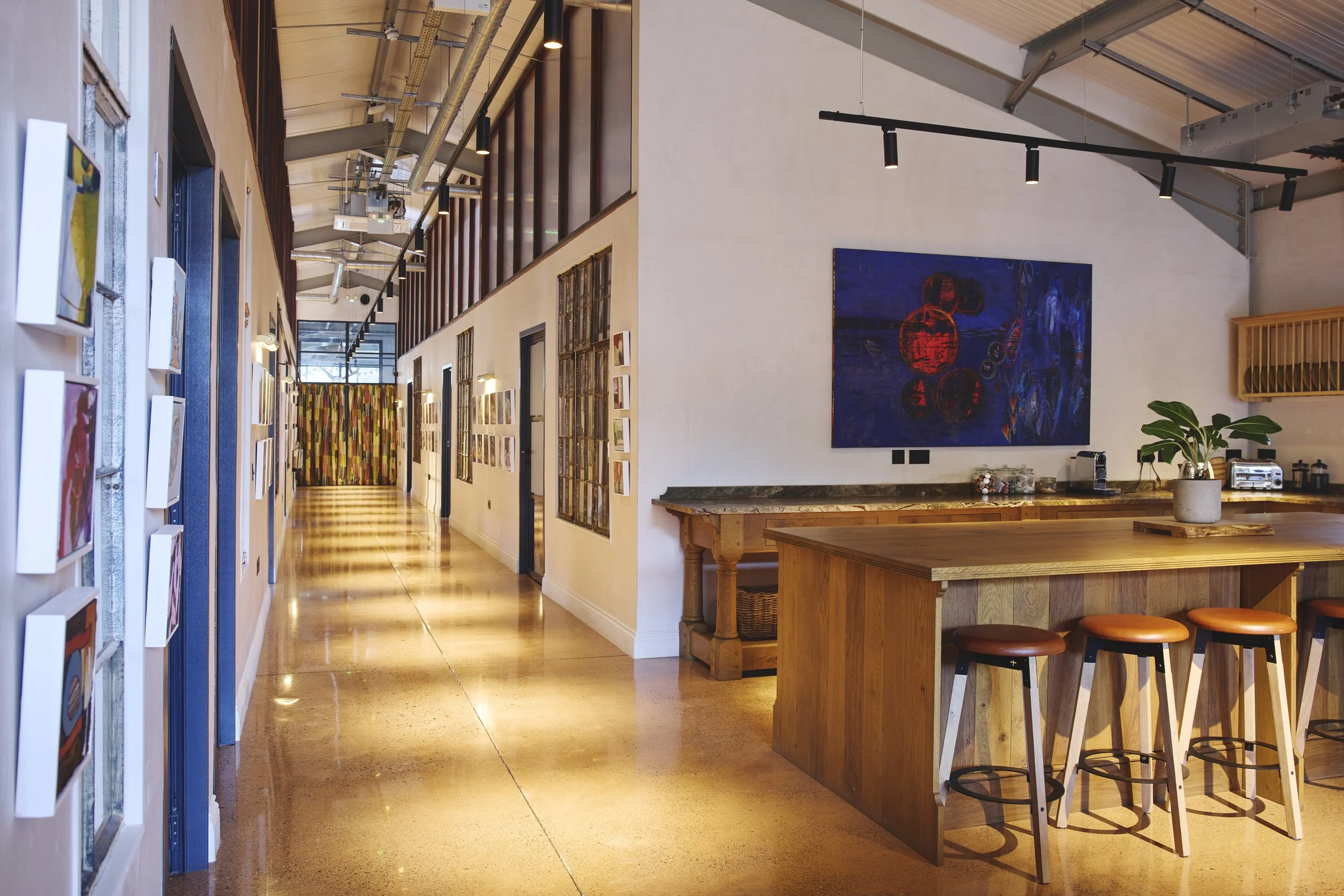 Interior of a modern art gallery or cafe with white walls, wooden furniture, and colorful abstract paintings on the walls. A hallway with a polished concrete floor leads to a colorful curtain at the end.