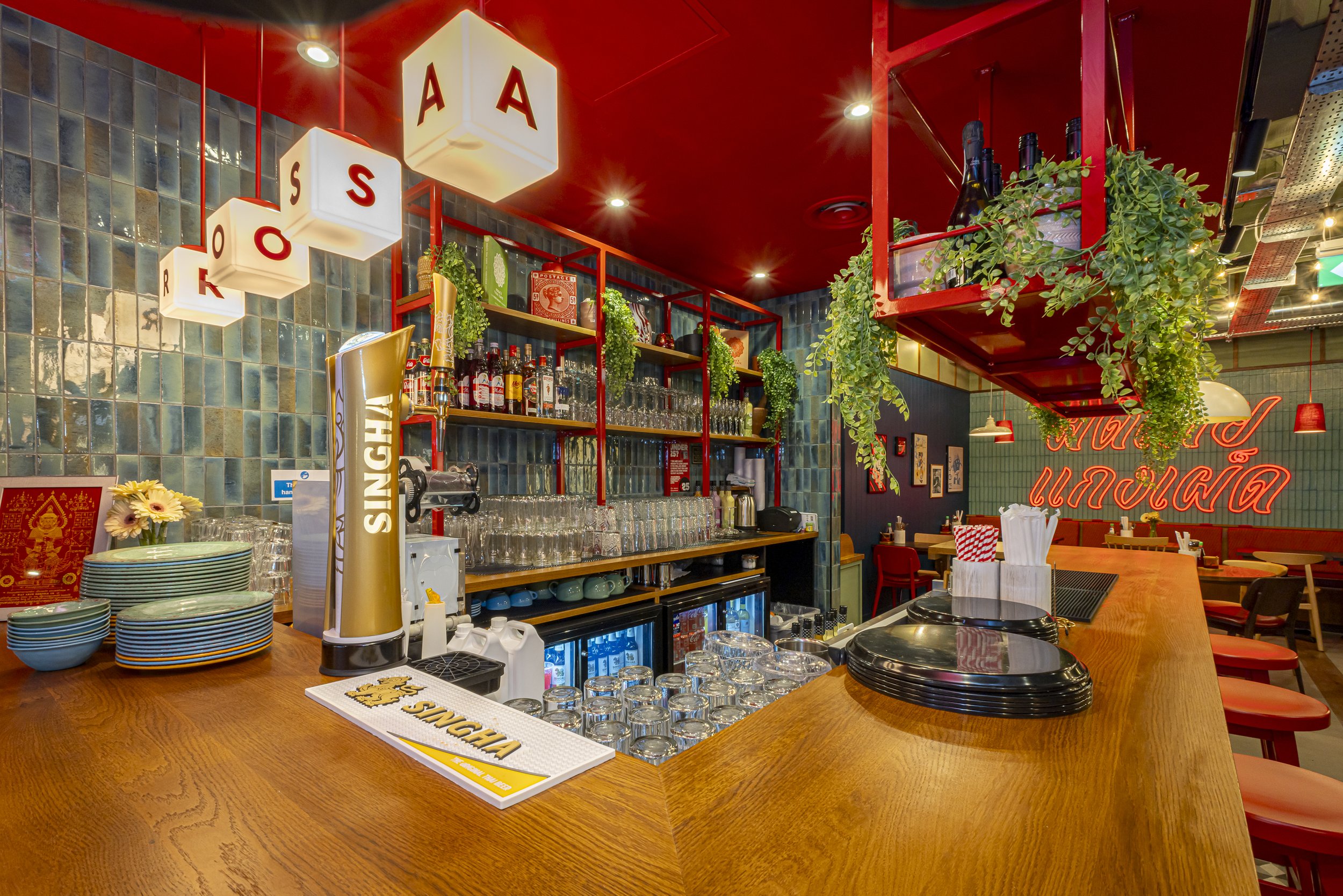 Inside a colorful bar or restaurant with a wooden counter, shelves stocked with bottles and glassware, hanging plants, decorative cubes with letters, and a neon sign in the background.