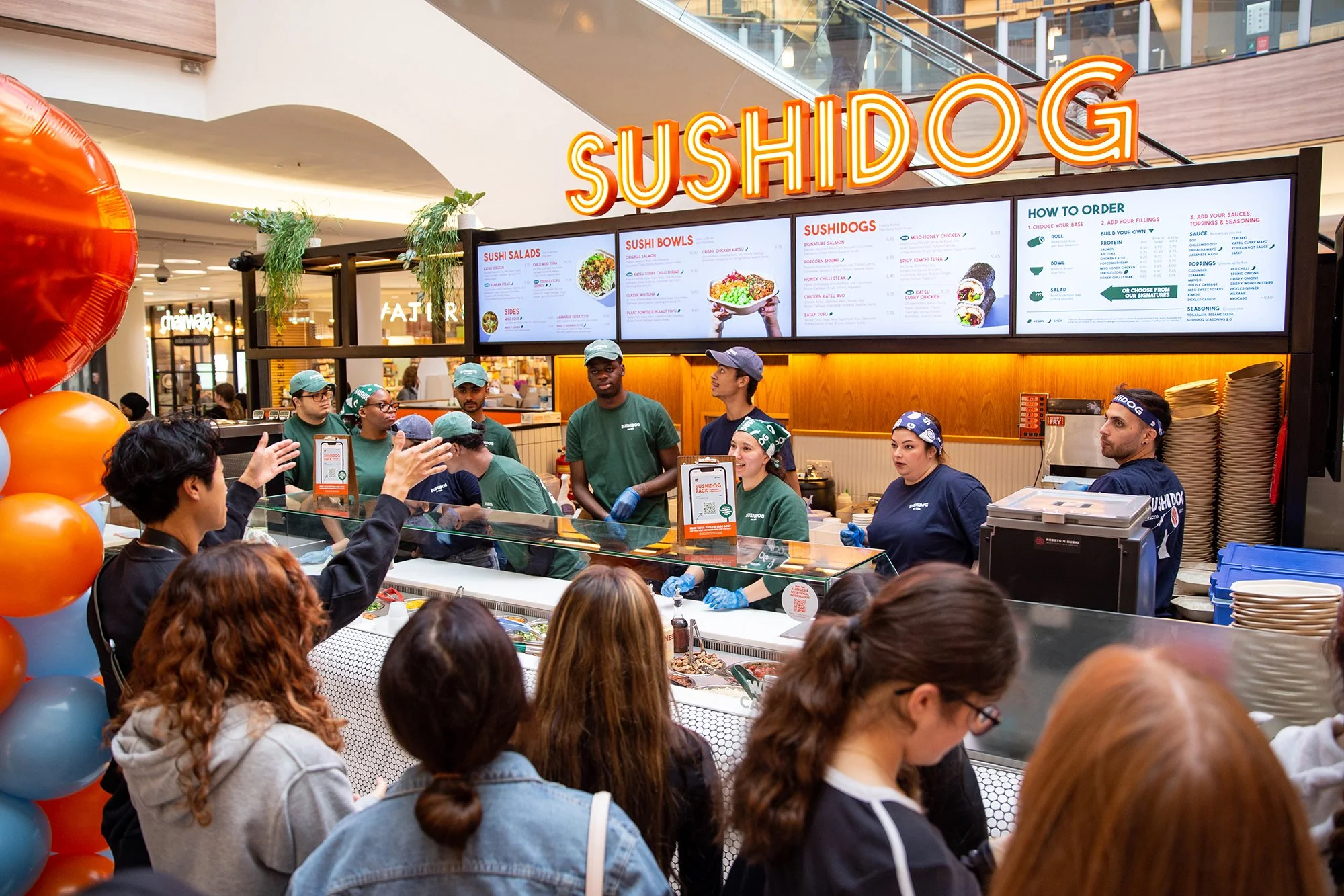 A busy sushi restaurant counter with staff preparing sushi and a group of customers waiting in line. The bright sign reads 'SUSHI DOG' and digital menu screens display sushi options and ordering instructions.