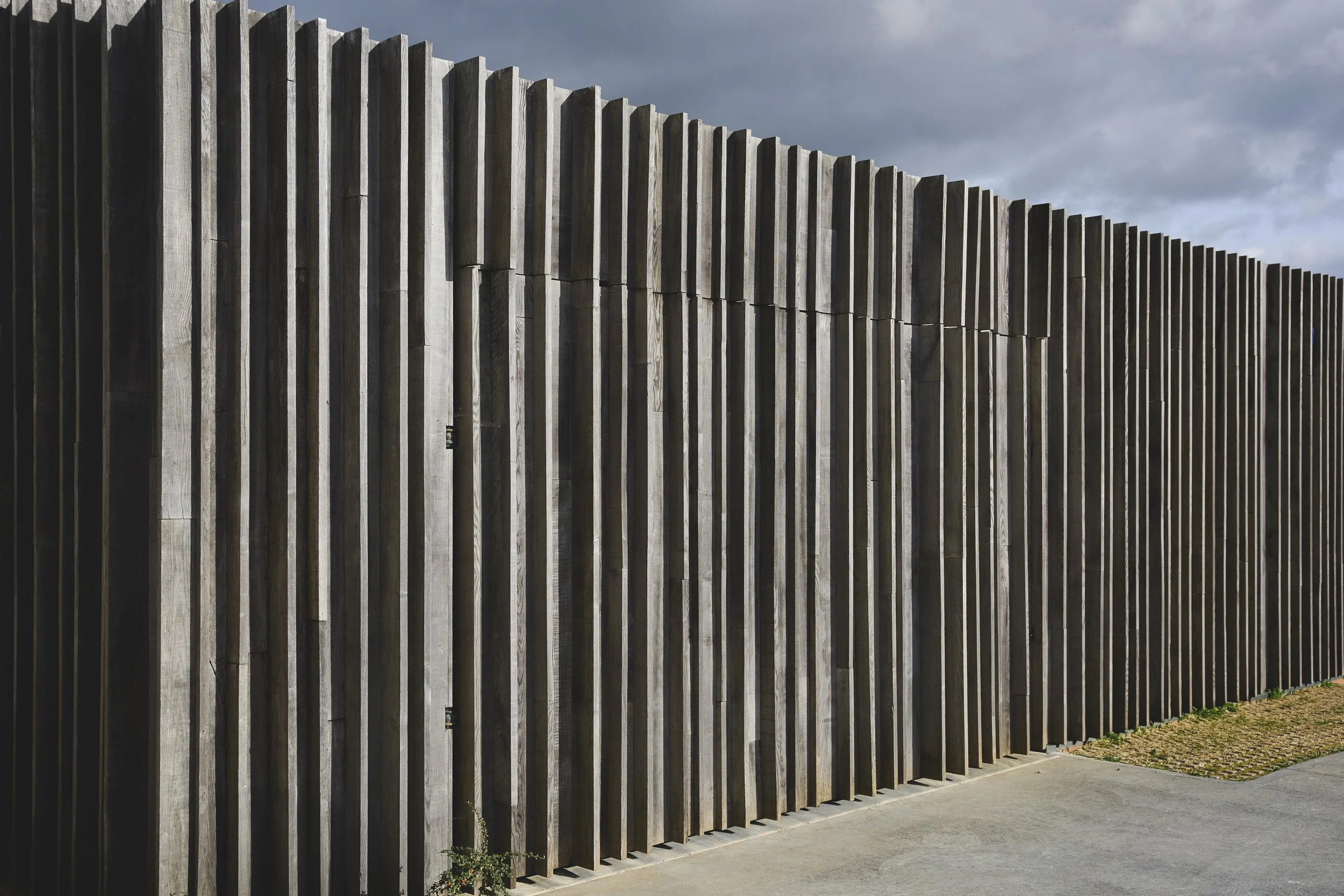A tall wooden fence with vertical slats under a cloudy sky, running along a paved surface.