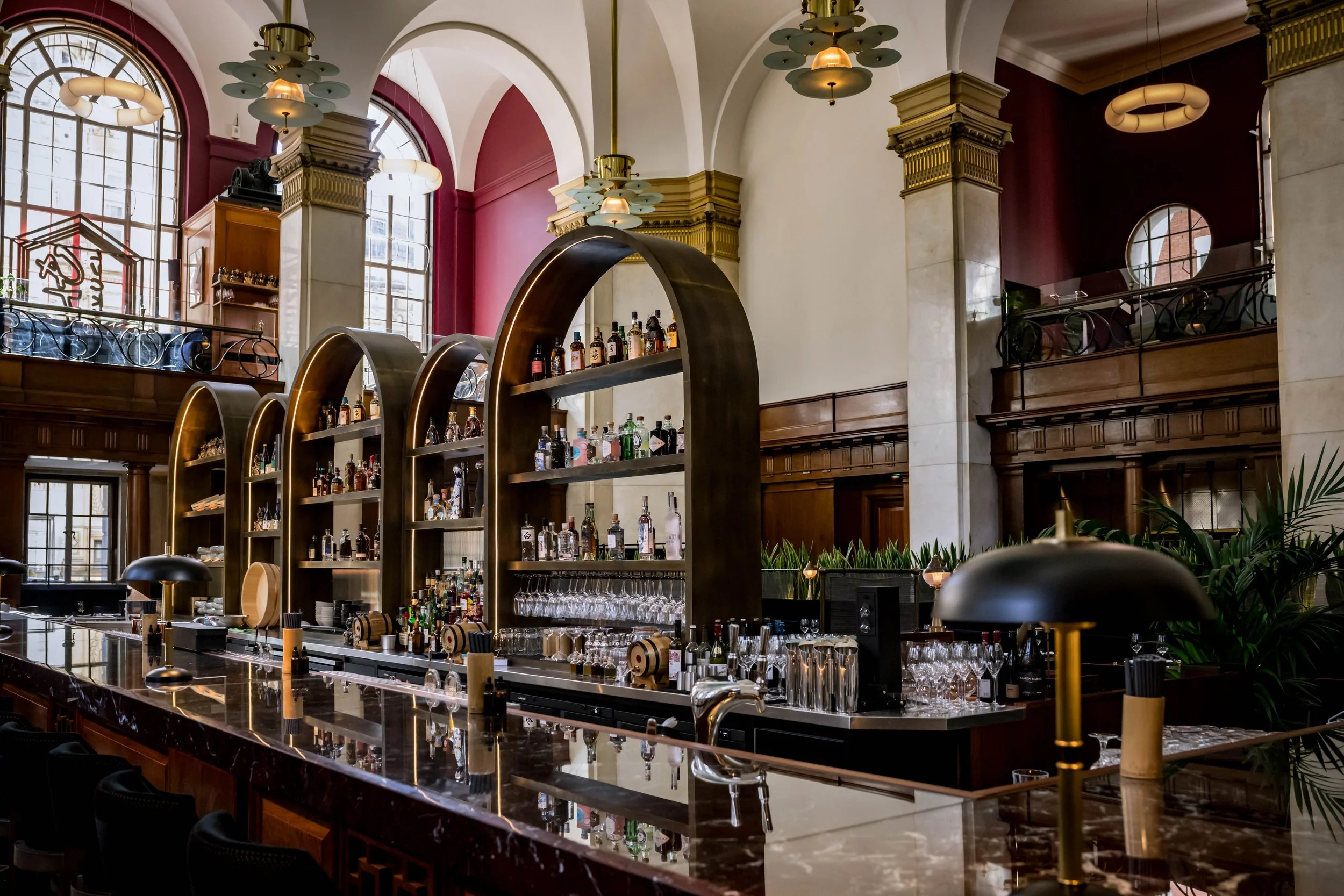Interior of a bar with a marble counter, modern black lamps, wooden shelves with alcohol bottles, and large windows in a high-ceilinged room with decorative columns and gold accents.