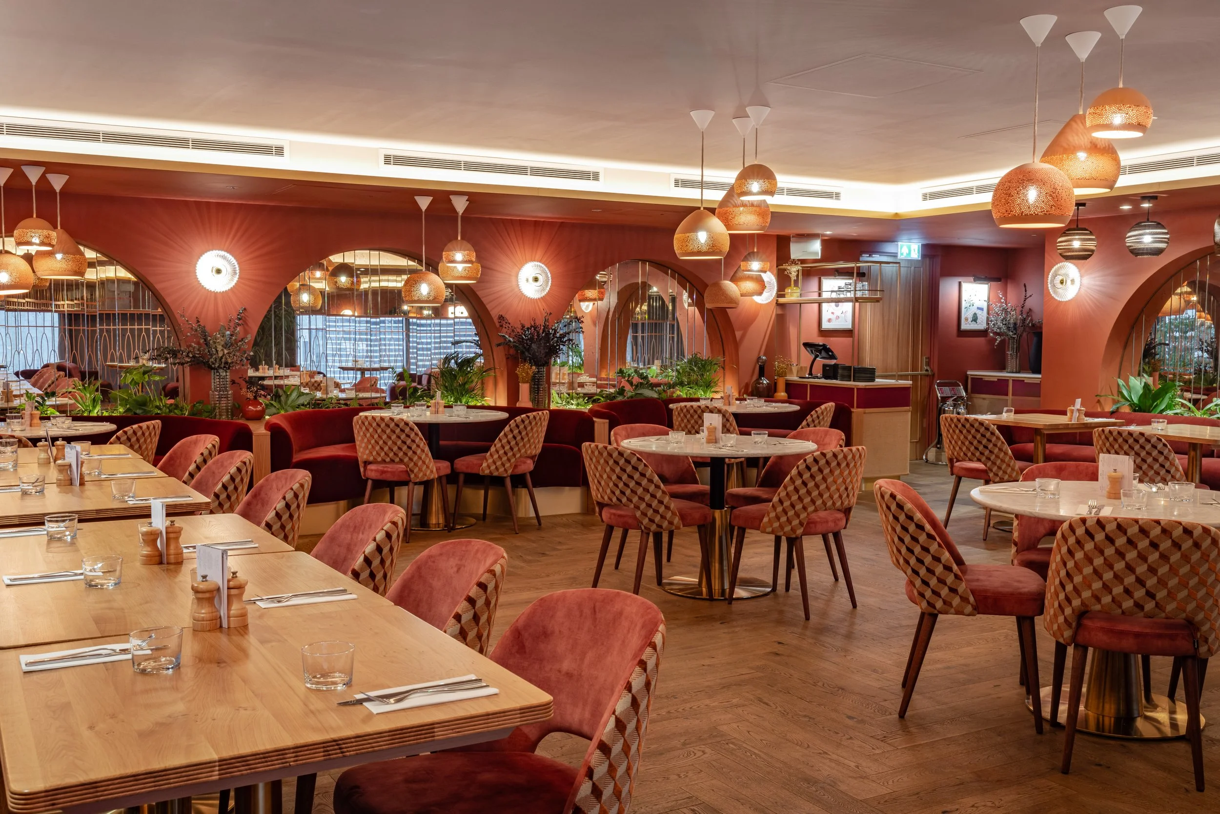 A cozy restaurant interior with pink walls, multiple tables set for dining, pink velvet and patterned chairs, hanging pendant lights, and decorative mirrors and plants.