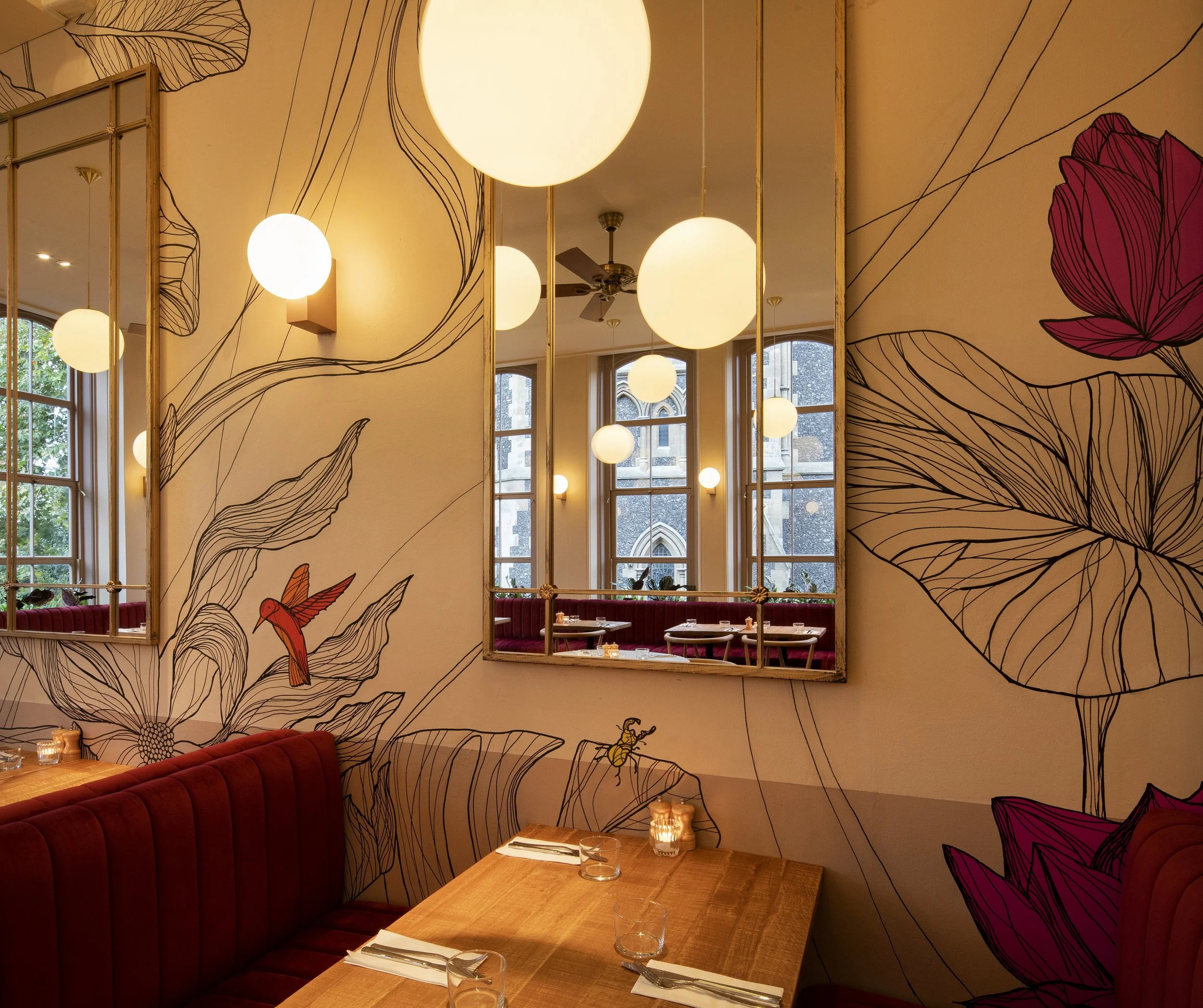 Interior of a restaurant with large mirrors on the wall, floral and bird wall art, red upholstered booths, wooden tables set with glasses and silverware, large circular pendant lights, windows showing an outdoor building and trees.