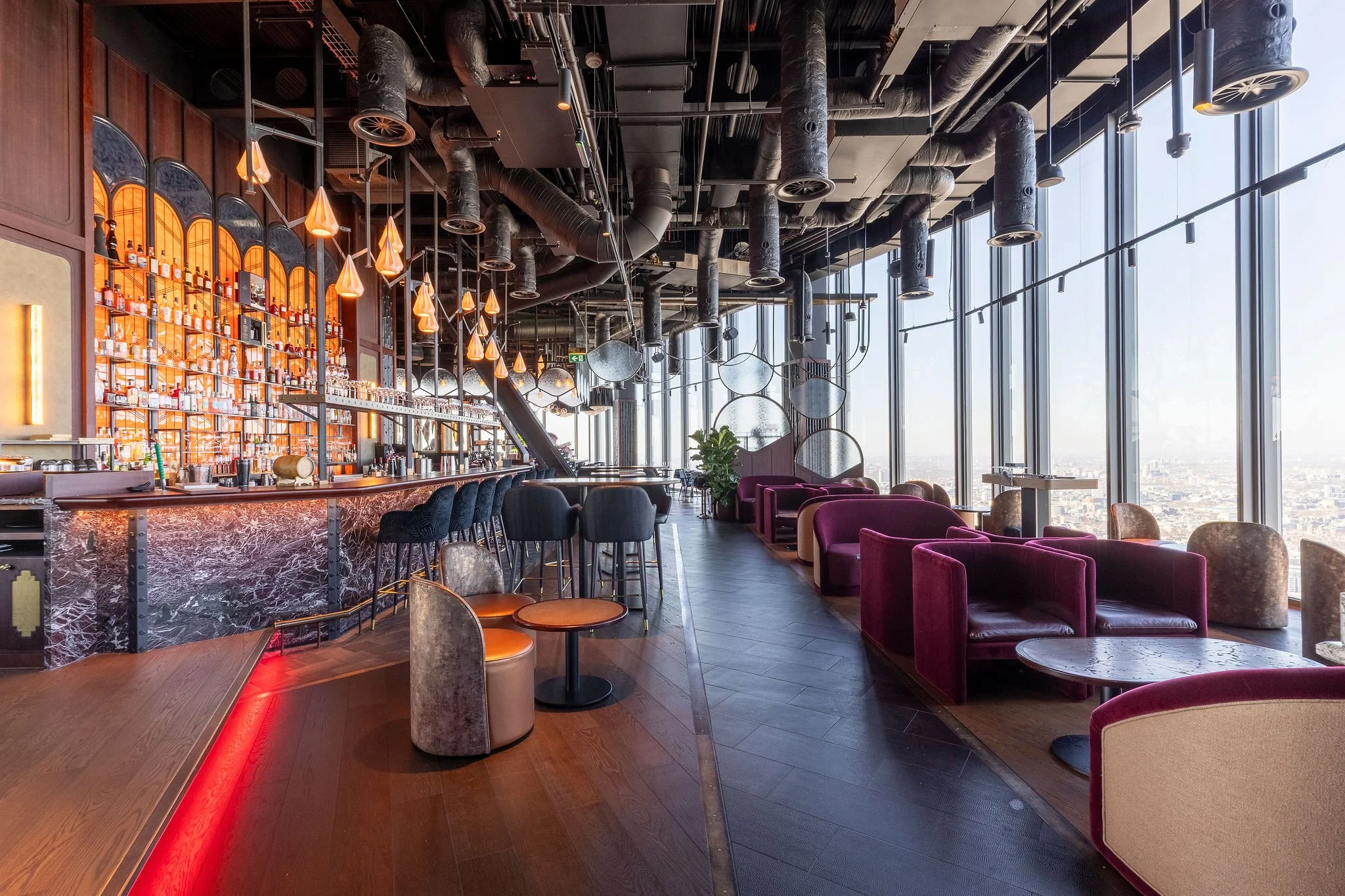 Interior of a modern lounge or bar area with large floor-to-ceiling windows, purple lounge chairs, a bar with barstools, and contemporary lighting fixtures with a city view.