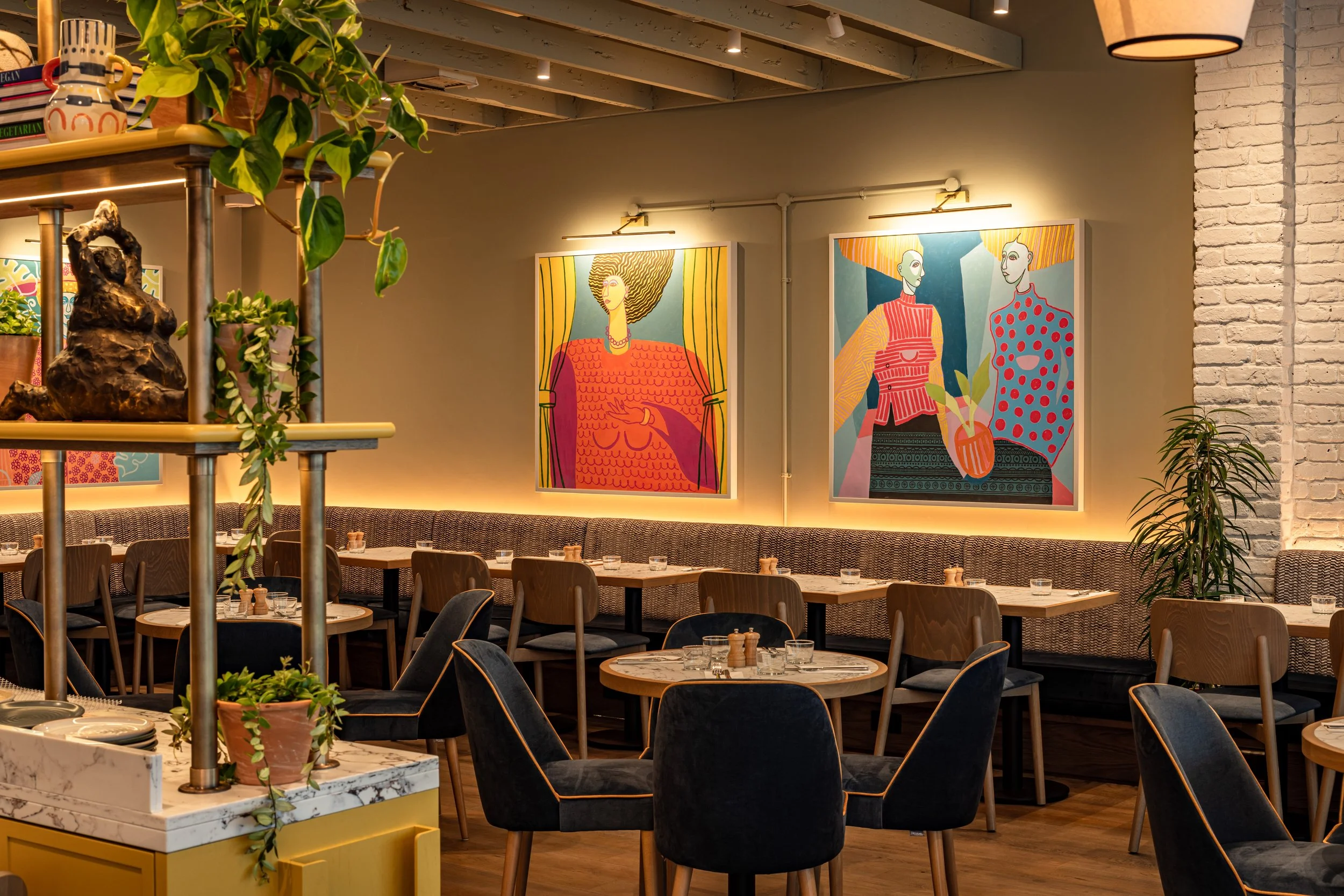Inside a modern restaurant with wooden tables and chairs, colorful artwork of women hung on the wall, potted plants, and warm lighting.