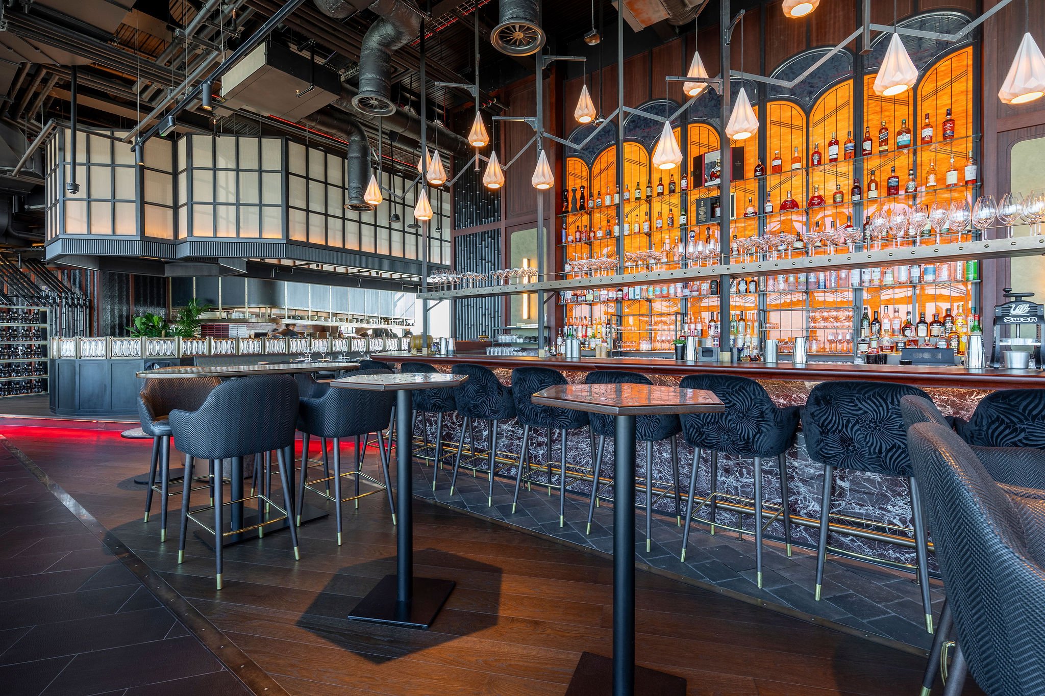 Modern upscale bar with a long leather-topped counter, high stools, and an extensive display of liquor bottles on backlit shelves. Contemporary pendant lighting hangs above, with wood and dark metal accents creating an elegant atmosphere.