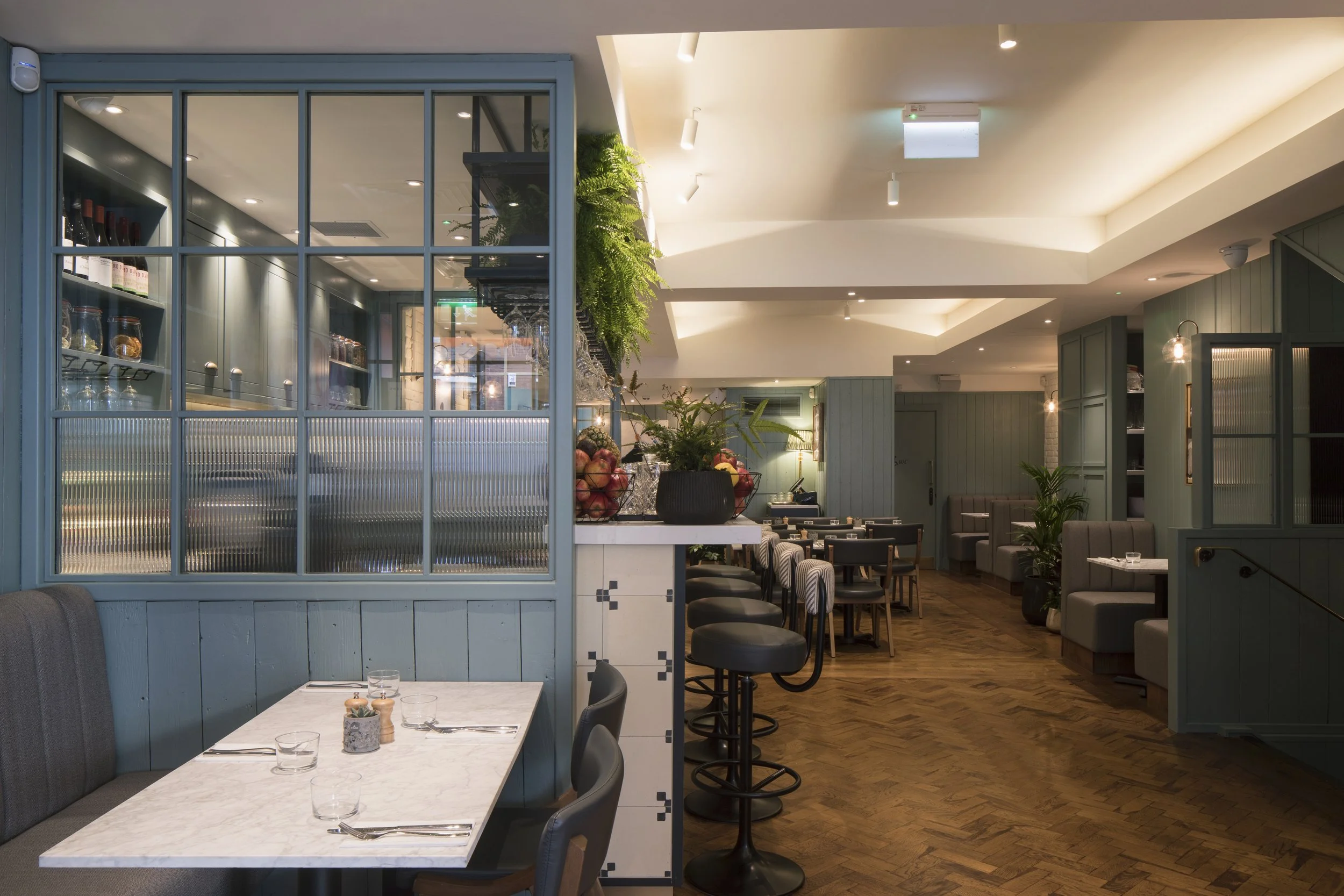Interior of a modern restaurant with soft lighting, a marble table set for dining, blue wall paneling, and various seating areas including booths and bar stools, complemented by green plants and decorative vases.