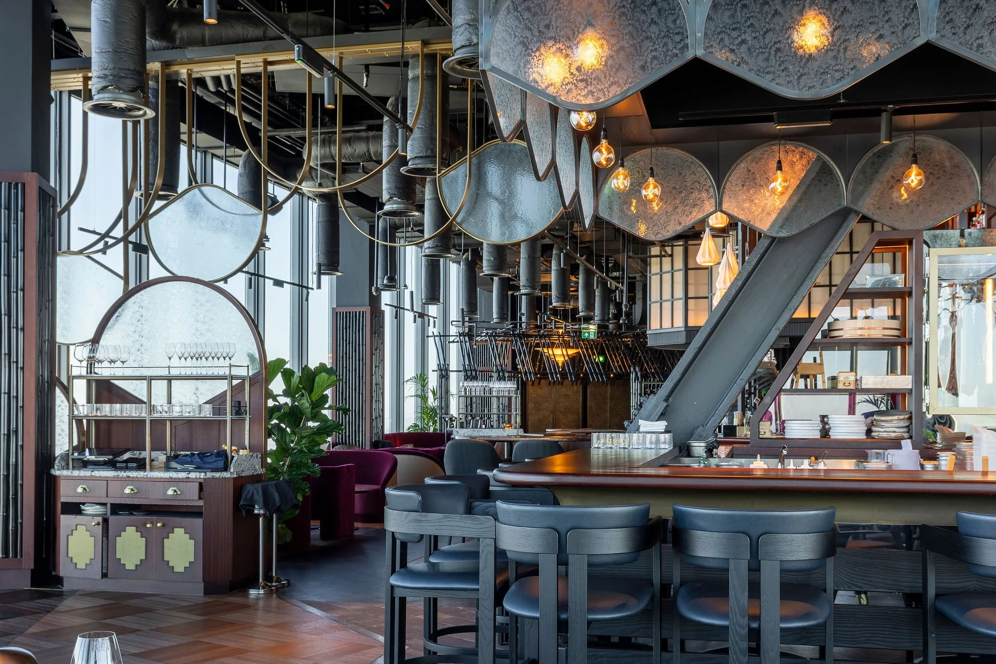 Interior of a modern restaurant or bar with a high ceiling, large windows, and artistic decorative elements hanging from the ceiling, including exposed light bulbs and metallic shapes. There is a curved bar counter with dark chairs, shelves with plat