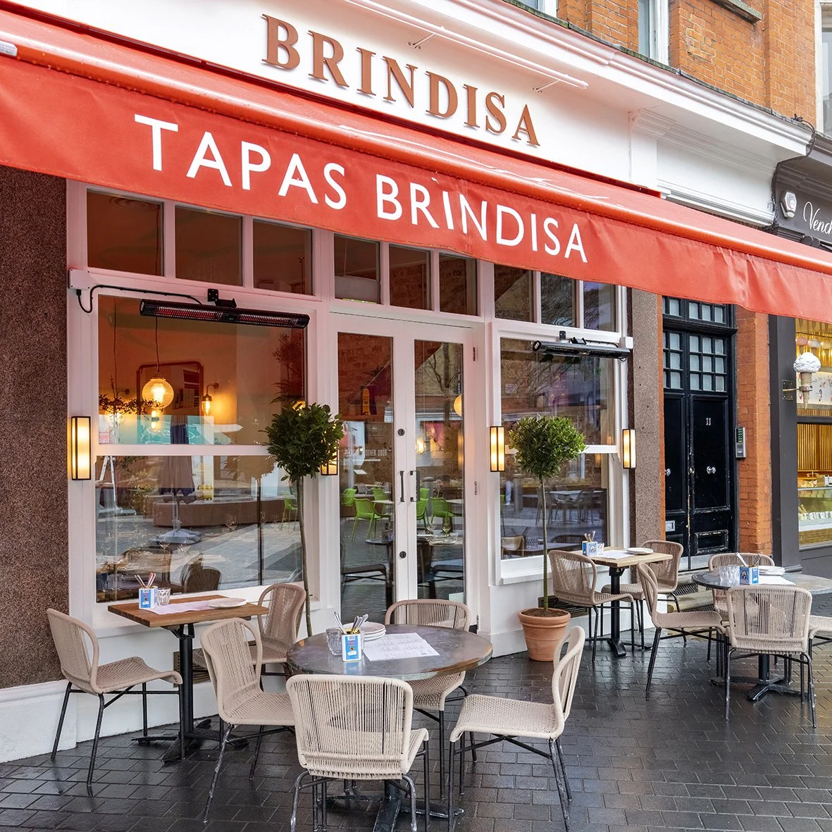 Exterior of a restaurant with a red awning that reads 'BRINDISA' and 'TAPAS BRINDISA'. The restaurant has glass doors and windows, outdoor seating with tables and chairs, and small potted trees.