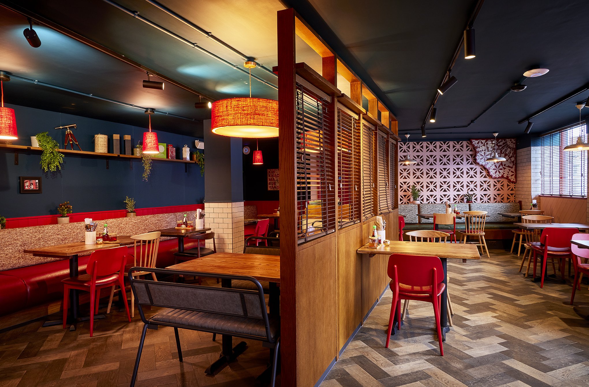 Interior of a cozy restaurant with wooden tables, red chairs, decorative shelves, hanging lamps, and patterned wall accents.