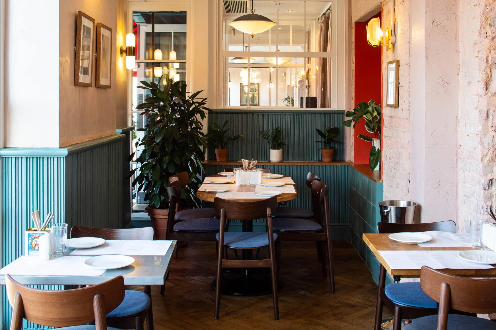 Interior of a cozy restaurant with tables set for dining, featuring wooden chairs around marble and wooden tables. Green plants line the back wall, and warm lighting fixtures hang from the ceiling. The space has a modern, inviting feel with light-colored walls and a mix of textures.