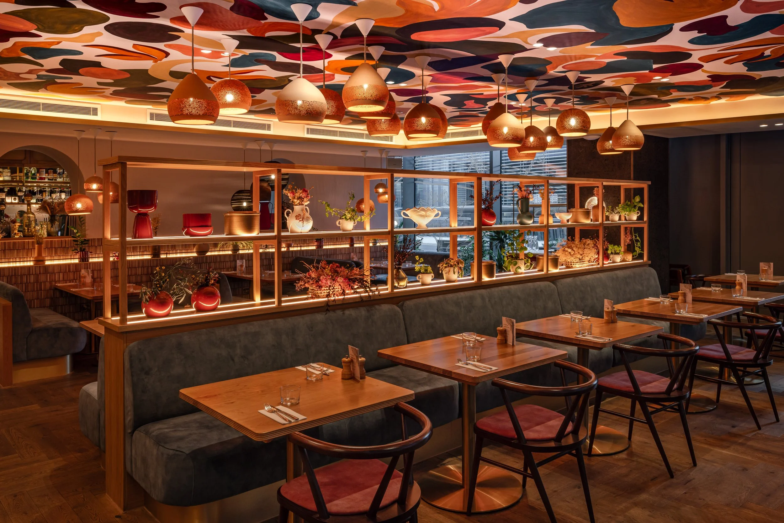 A cozy restaurant interior with wooden tables and chairs, decorative hanging lights, and a shelf filled with plants and vases, illuminated by warm lighting.