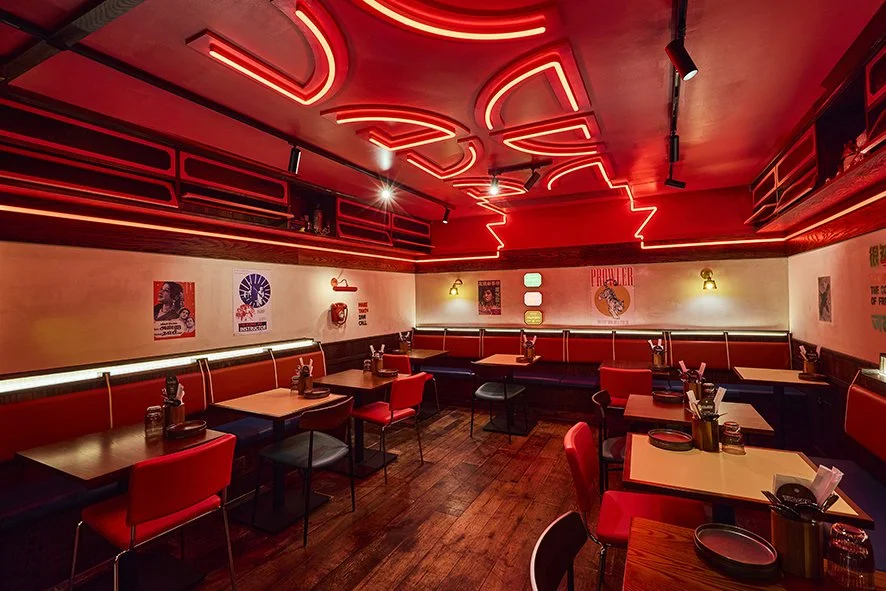 A dimly lit restaurant or club with red neon lighting on the ceiling and walls, red chairs, and tables set with plates, utensils, and condiments. The walls have posters and decorative lighting.