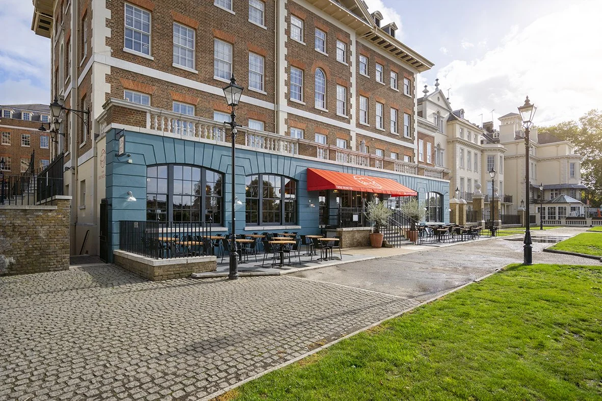 Exterior view of a restaurant with outdoor seating, surrounded by historic buildings and street lamps, on a sunny day.