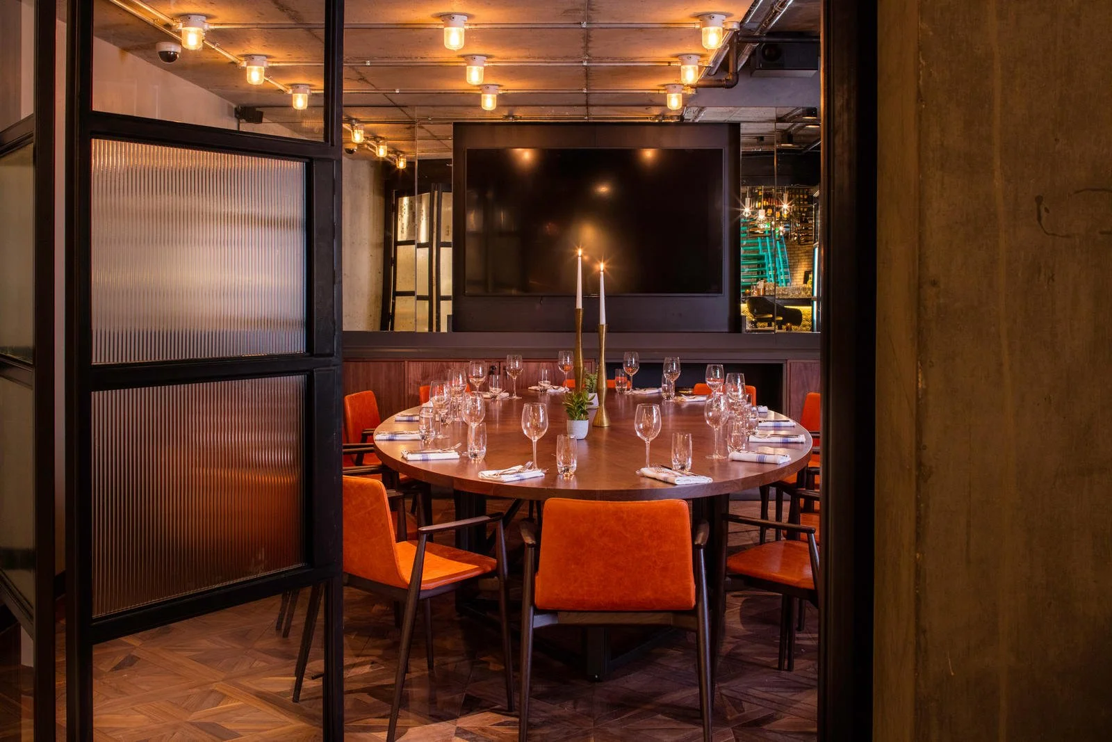 Elegant restaurant dining room with a large round wooden table set with wine glasses, plates, napkins, and a centerpiece with two tall candles. The room features warm lighting, modern decor, and a large dark television screen on the back wall.