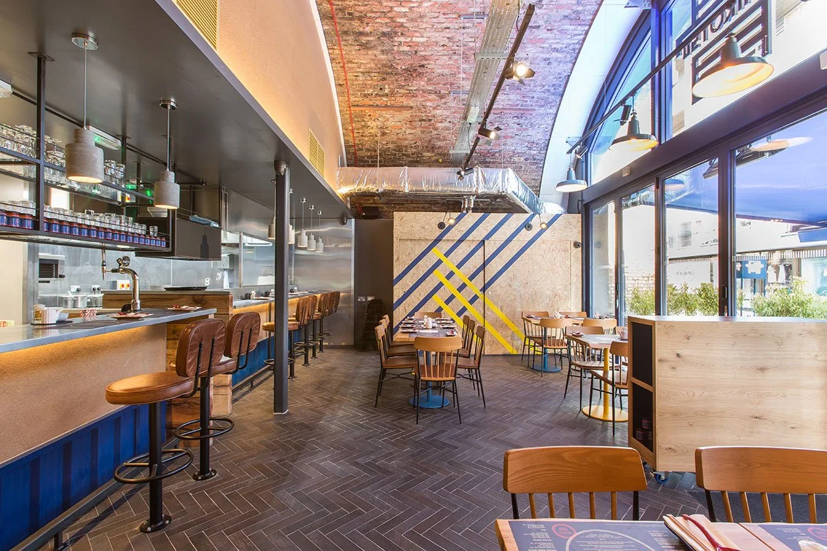 Empty modern restaurant with a bar on the left side, high chairs, and tables set for dining near large windows with blue shades, featuring a brick ceiling and contemporary wall art.