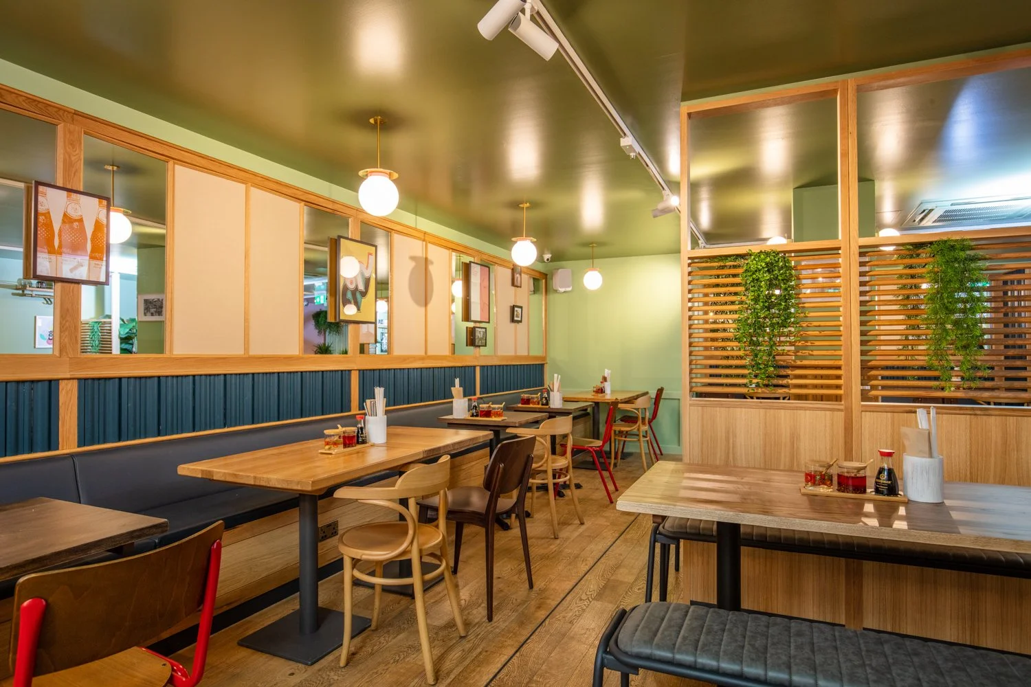 Empty restaurant interior with wooden tables, mixed chairs, green walls, decorative art, partition with greenery, and warm lighting.