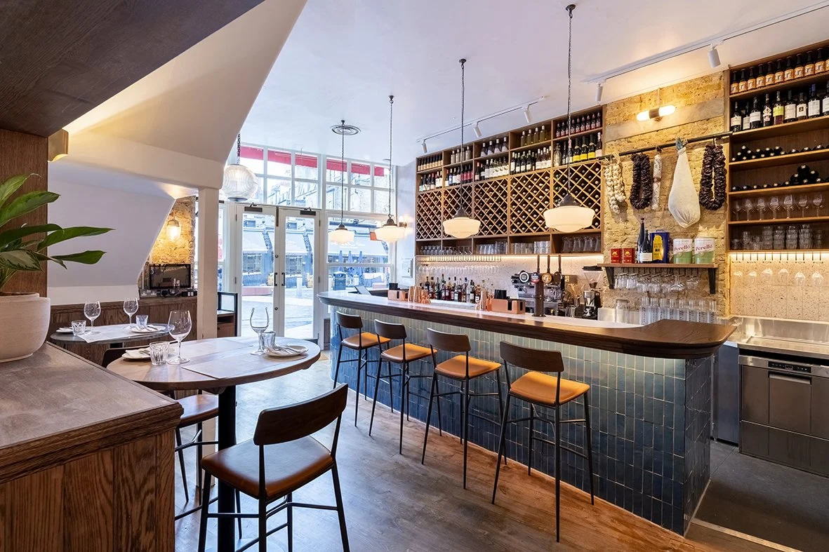 Interior of a modern restaurant or bar with a curved bar counter, bar stools, shelves with bottles and glasses, and a windowed entrance showing a street outside.