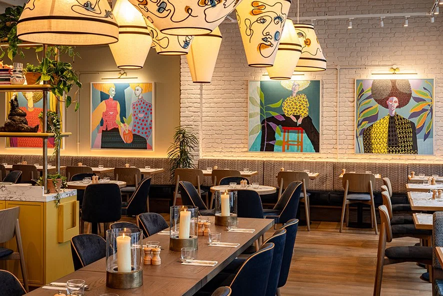 Interior of a modern restaurant with artistic portraits on the brick wall, hanging decorative lamps, and tables set with candles and glassware.