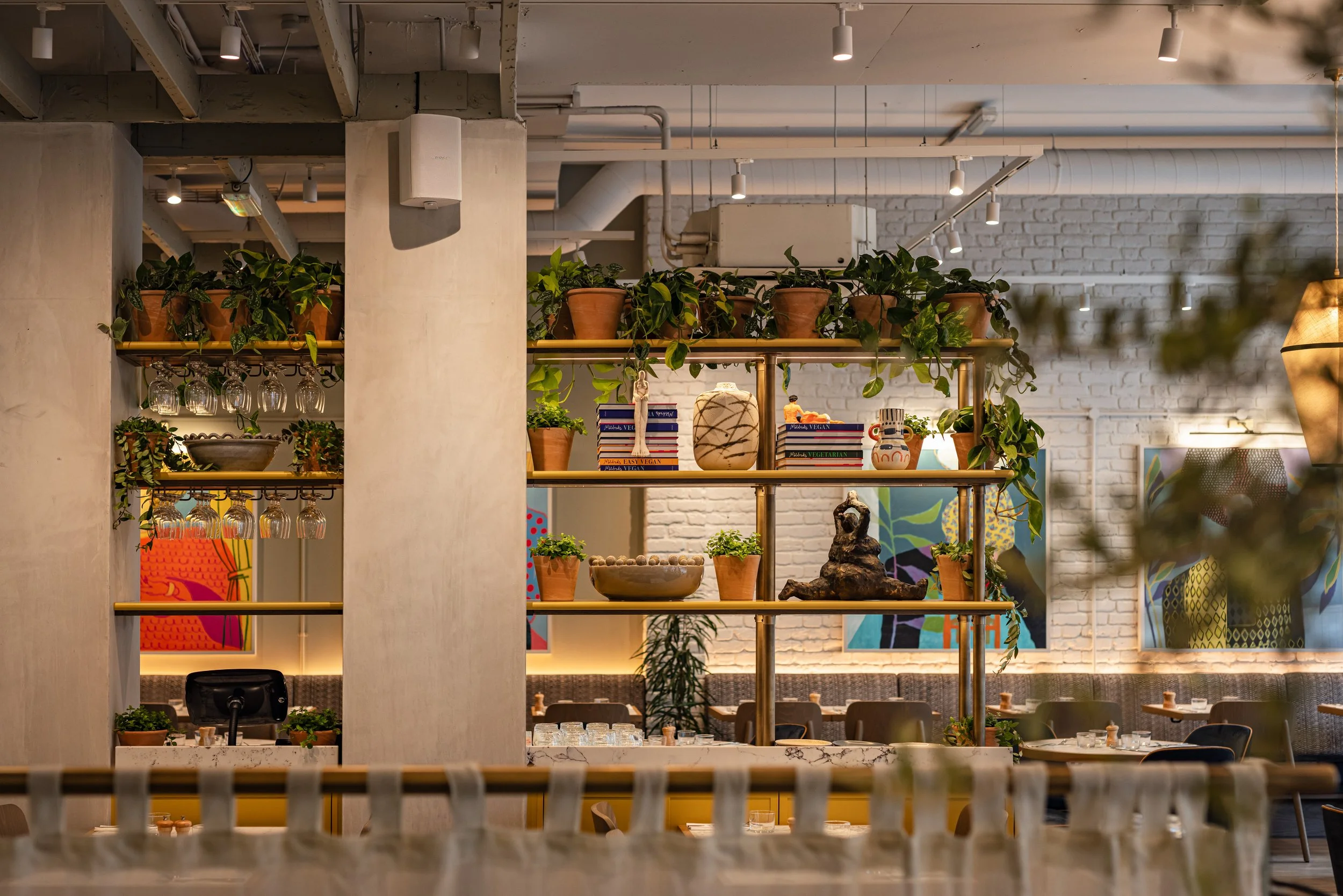 Interior of a restaurant with a decorative shelving unit holding potted plants, books, and art pieces, with colorful paintings on the brick wall behind.