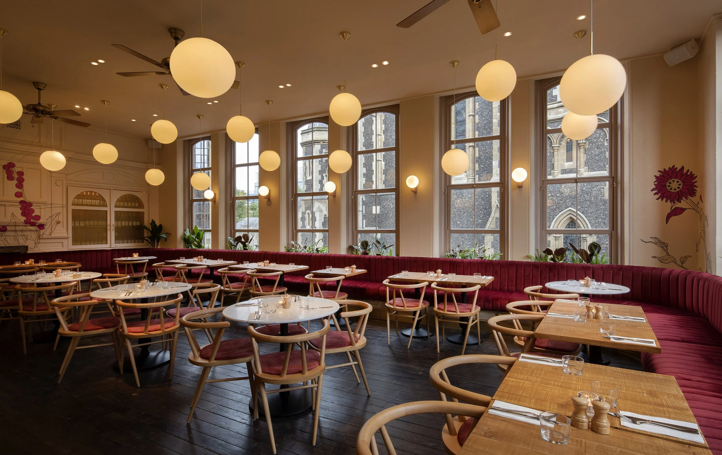 Empty restaurant with round and rectangular wooden tables, pink chairs, a long red cushioned banquette, large windows, hanging spherical lights, and a view of a stone church outside.