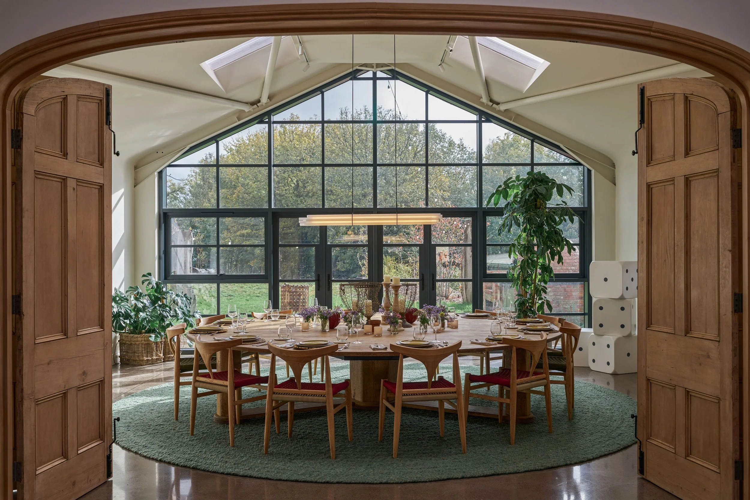 A dining room with a round wooden table set with plates, glasses, and candles, surrounded by wooden chairs with red cushions. The room features large windows with a view of the outdoors, two potted plants, and a side stack of oversized dice. The ceil