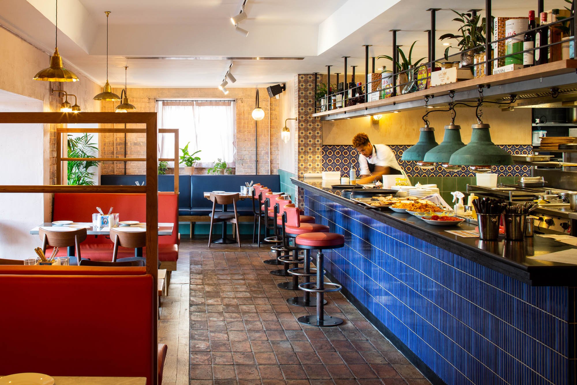 Interior of a modern restaurant with a bar counter, seating area with tables and chairs, decorative plants, and a staff member preparing food behind the bar.