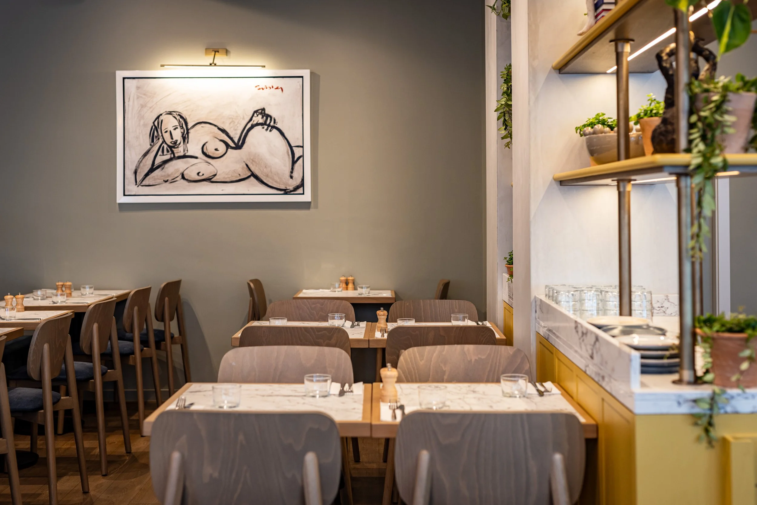 Interior of a modern restaurant with wooden tables and chairs, a large framed abstract art piece on the wall, and a shelf with potted plants and glasses.