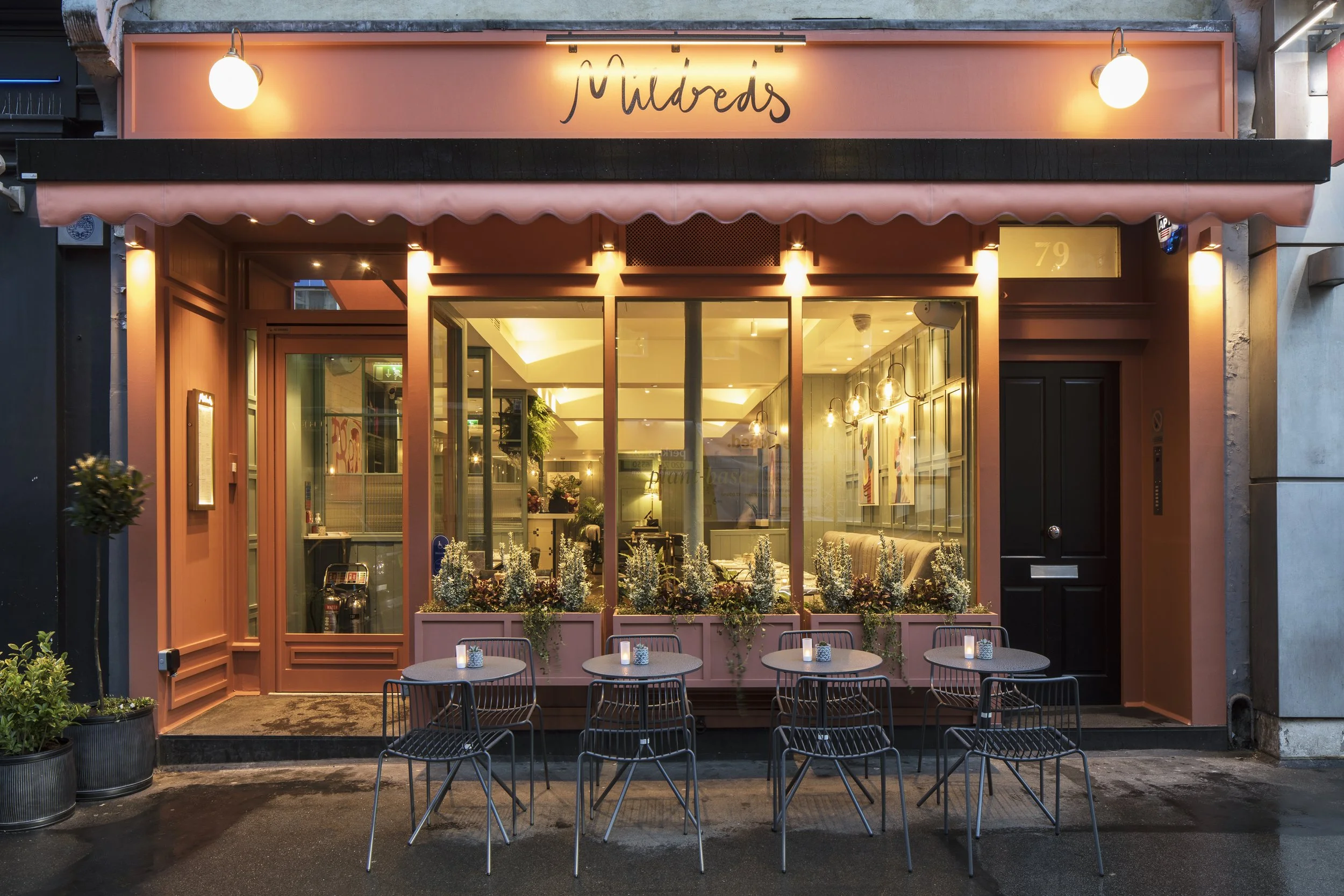 Pink storefront with outdoor seating and large front windows, sign reading 'Mildreds' above, illuminated by exterior lights, with four tables and chairs outside.