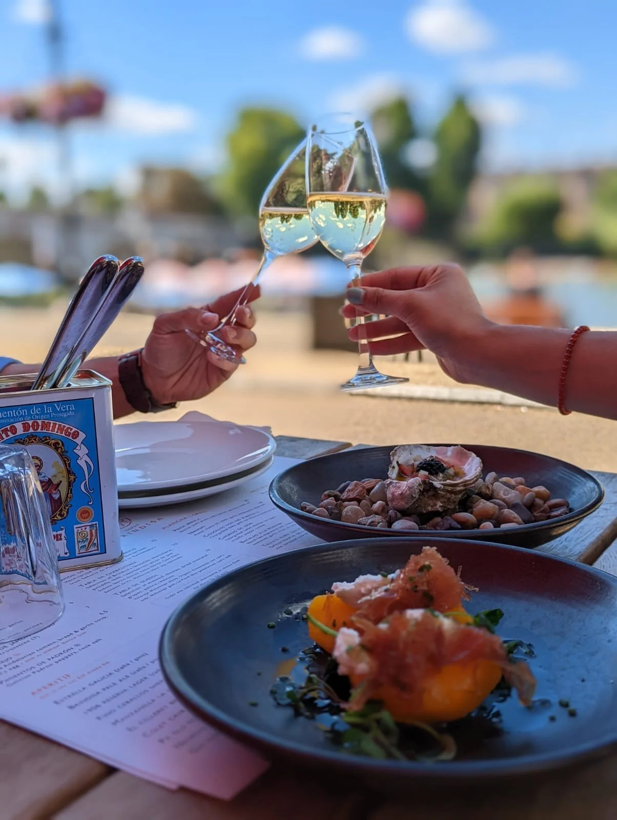 Two people clinking glasses of white wine at an outdoor restaurant table with plates of food, including a salad with peaches and prosciutto, and an oyster, with a river and trees in the background.