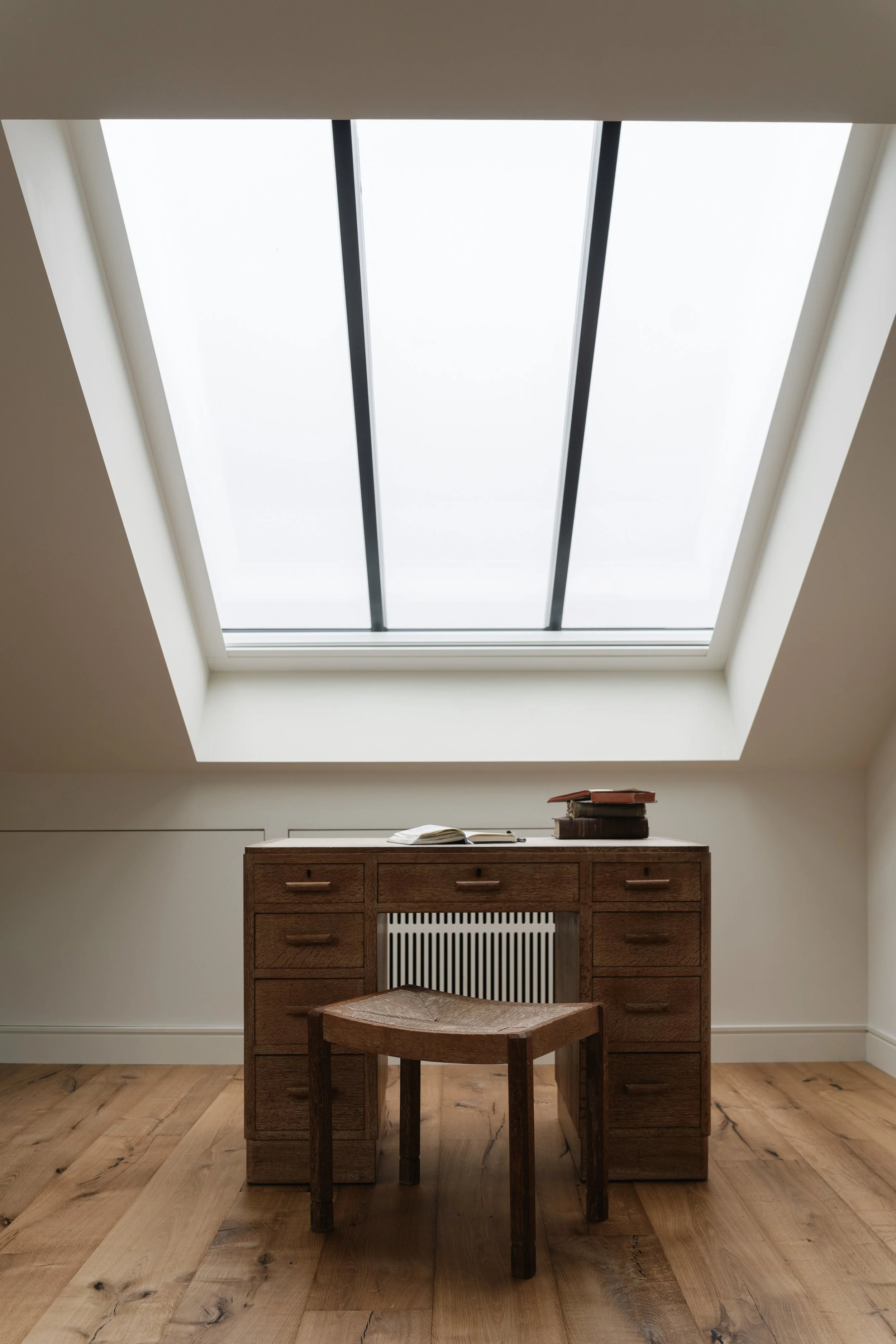 A wooden desk with drawers and a chair in front of a radiating heater beneath a skylight window in an attic room.