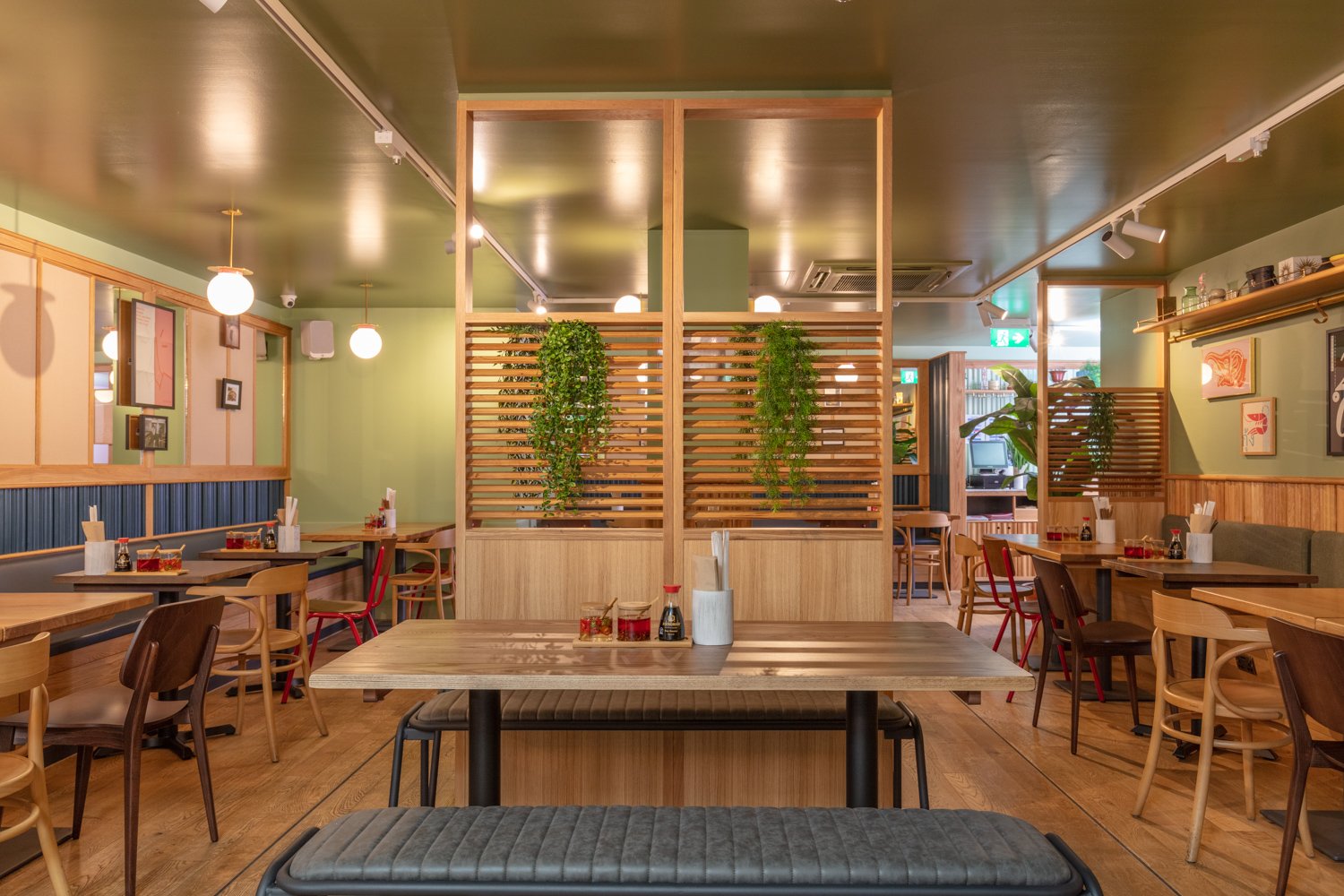 Interior of a restaurant with wooden tables and chairs, decorated with green plants, framed artwork, and soft ceiling lighting.