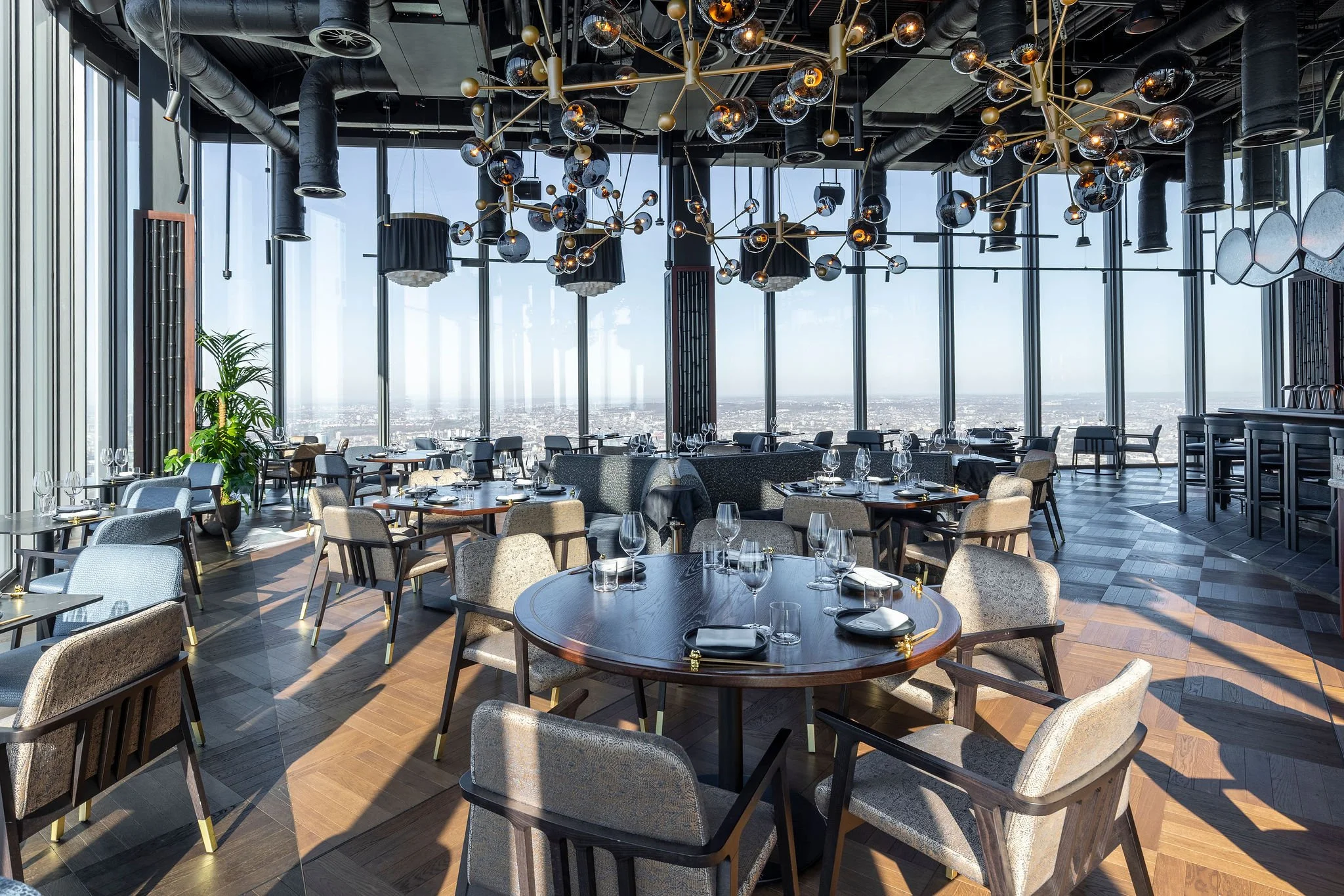 Modern upscale restaurant with floor-to-ceiling windows, contemporary chandeliers, round tables set with wine glasses, black cloth napkins, and silverware, and a view of the city skyline.
