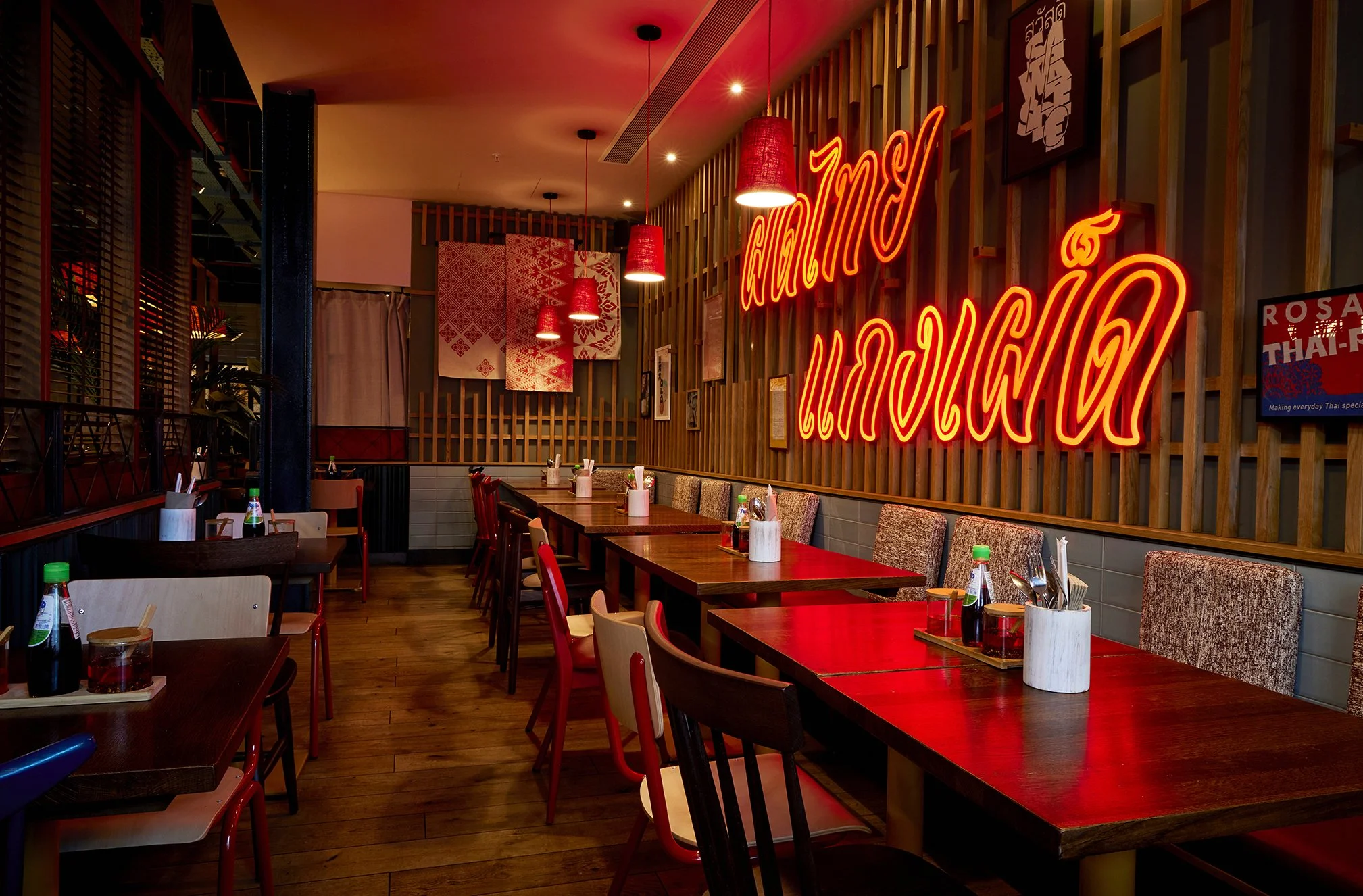 Interior of a restaurant with wooden tables, chairs, and red lighting. There is a bright red neon sign in Thai script on the wall, along with hanging red pendant lights and decorative wall art.