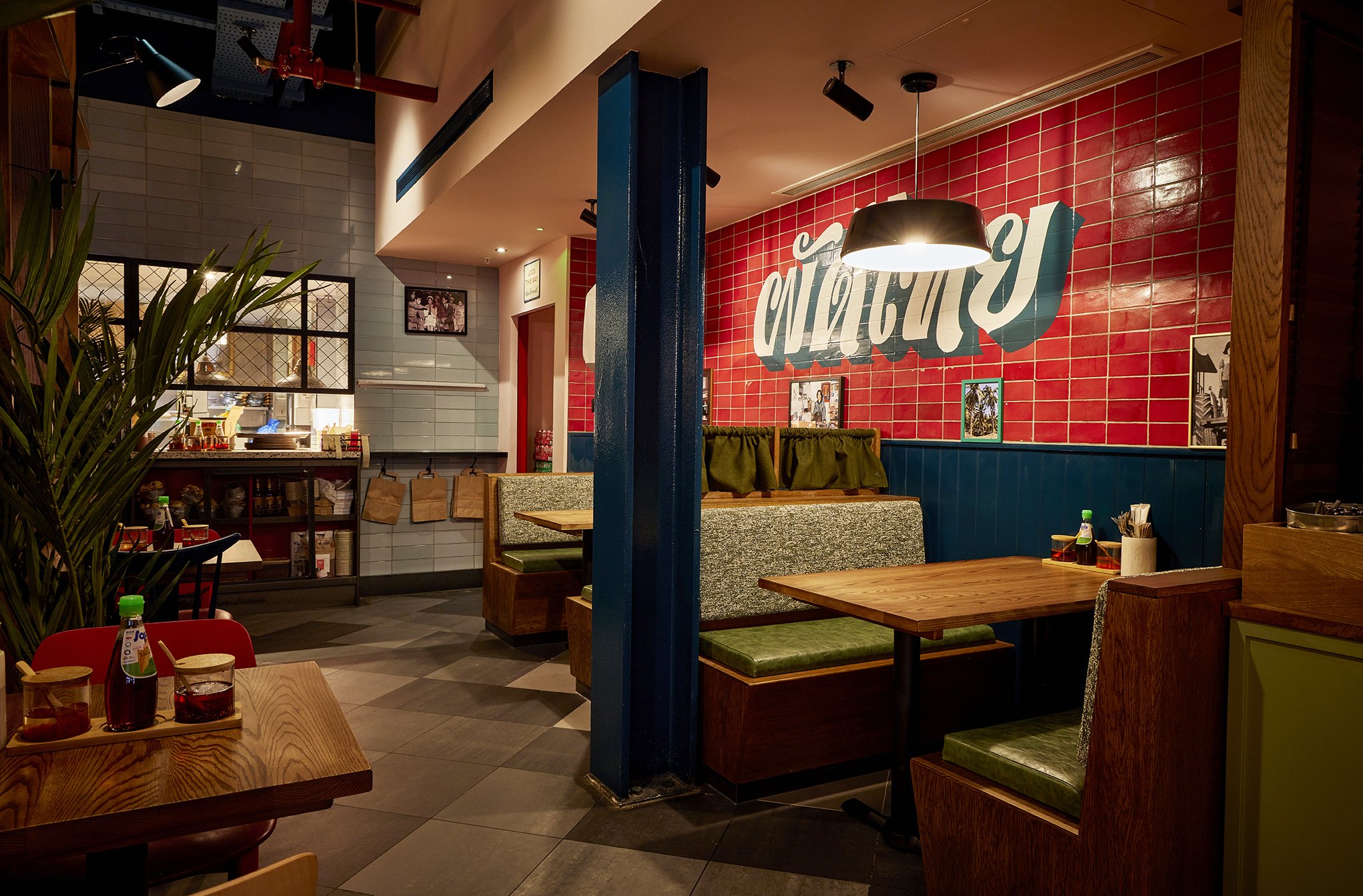 Interior of a cozy restaurant with wooden tables and green cushioned seats, a red tiled wall with white and teal signage, and a partially visible open kitchen area in the background.