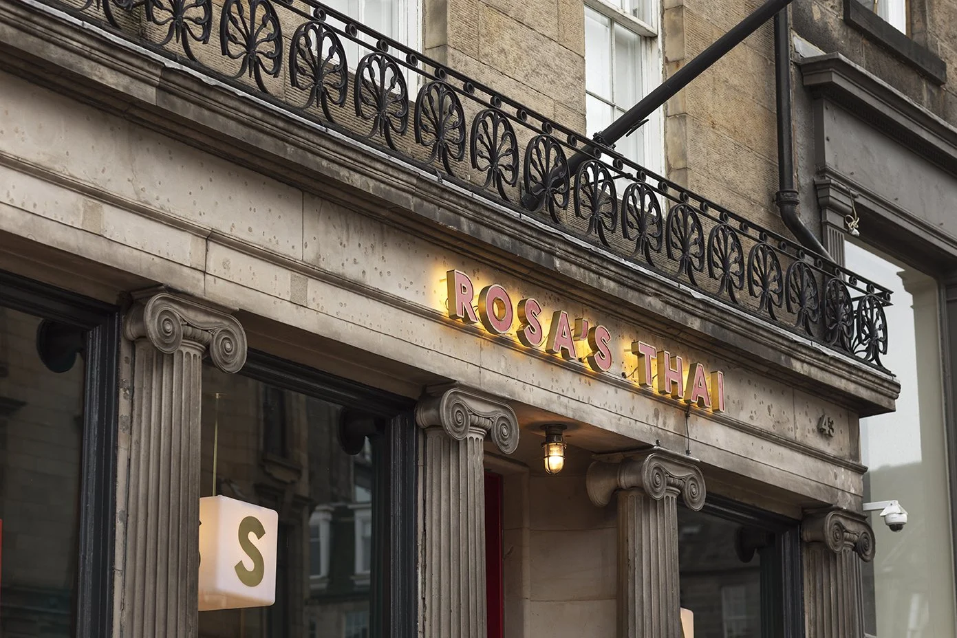 Exterior of a building with a sign that reads 'Rosa's Thai' in pink neon letters. The building has classical architectural details including columns with Ionic capitals and a decorative metal balcony.