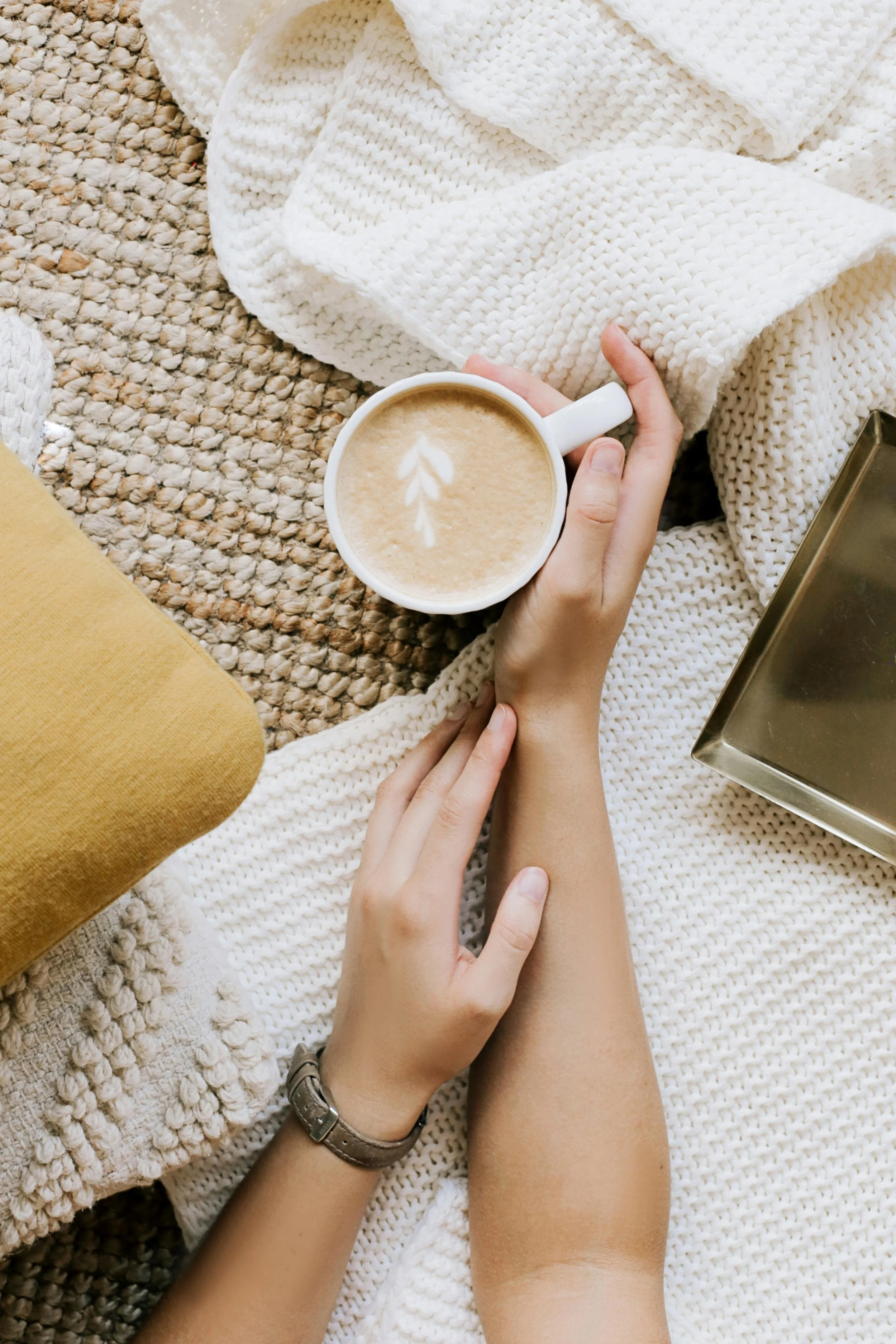 Person holding a mug of coffee with latte art, resting on a textured blanket with a yellow pillow and a metal tray nearby.