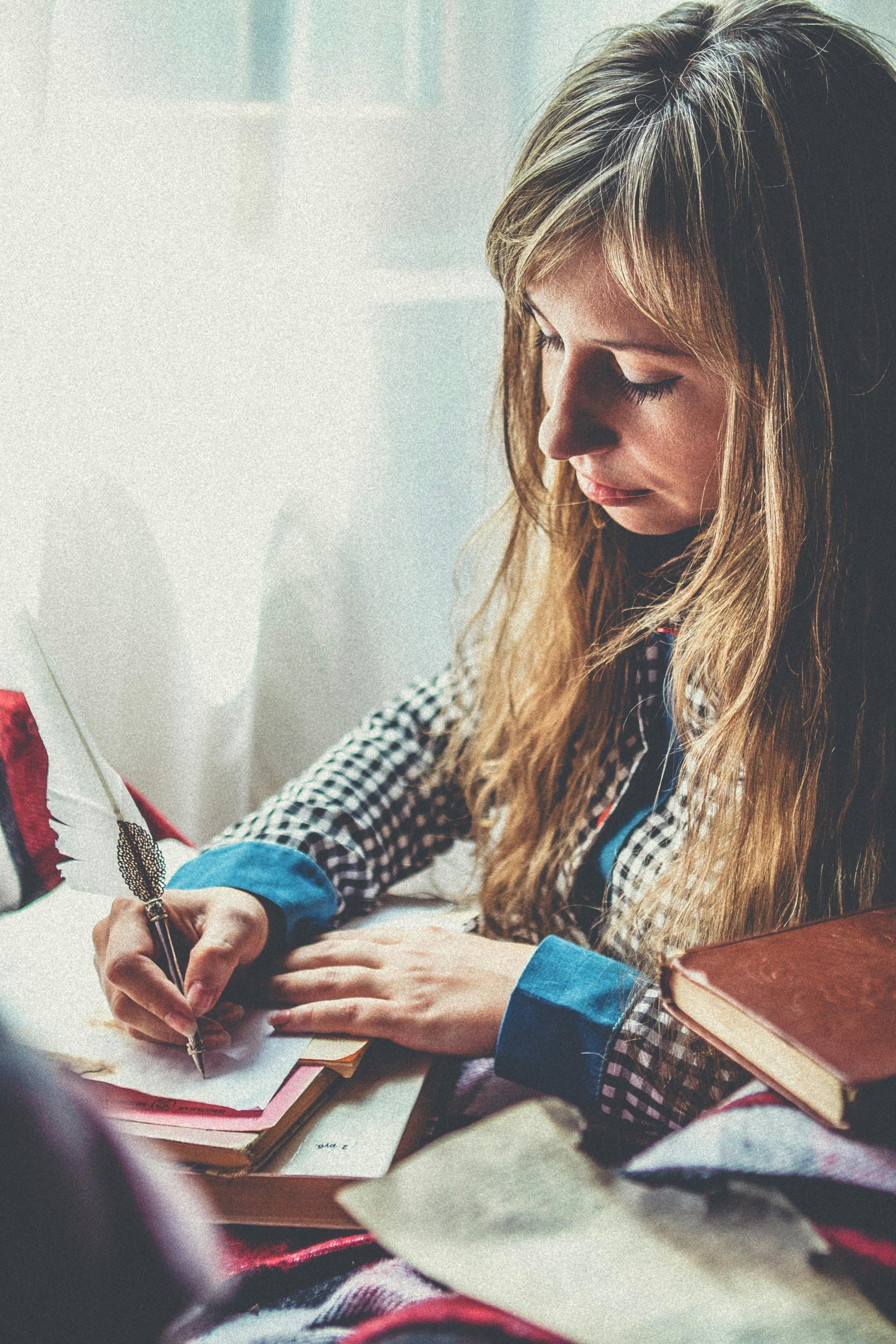 A woman with long blonde hair and a checkered shirt is writing with a decorative feather pen on paper at a desk. There are books and papers around her.