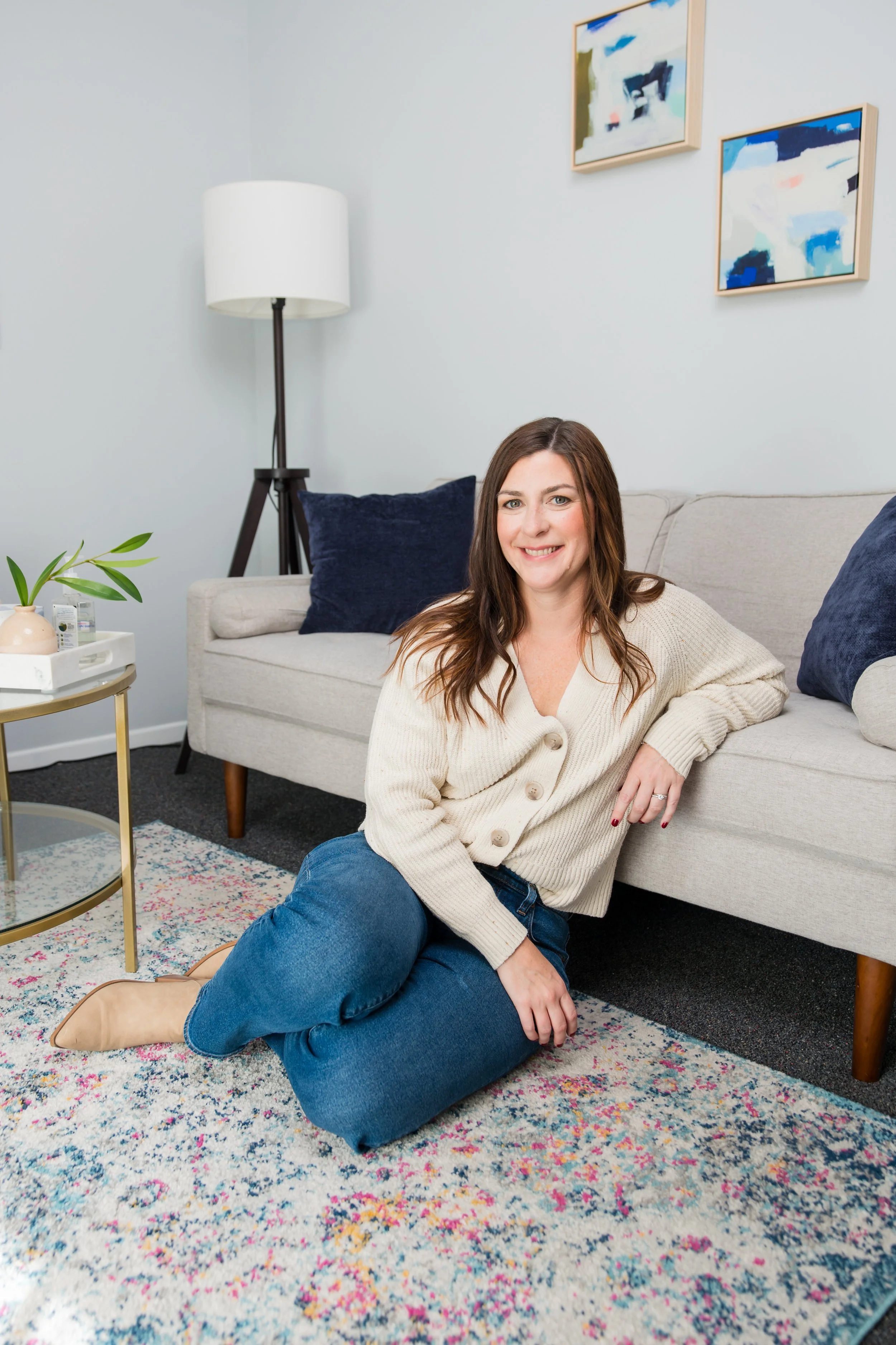 A woman with brown hair sitting on a colorful rug in a living room, smiling at the camera. She is wearing a cream sweater and blue jeans, with cream boots. Behind her is a light-colored sofa with dark blue pillows, a potted plant on a gold table, and a white lamp. There are framed abstract art pieces on the wall.
