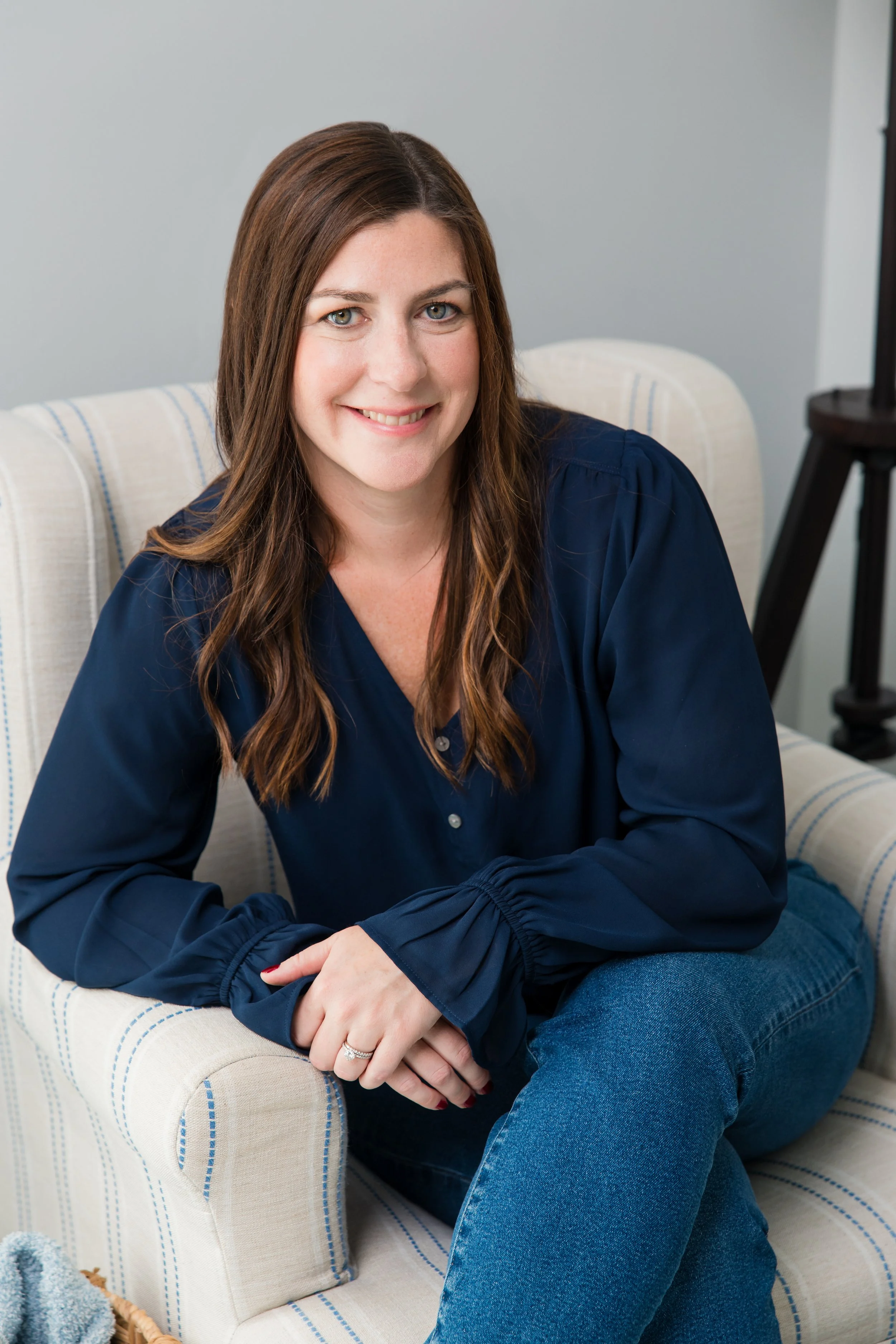 A woman with long brown hair, wearing a navy blue blouse, seated on a light-colored armchair, smiling at the camera in a cozy room.