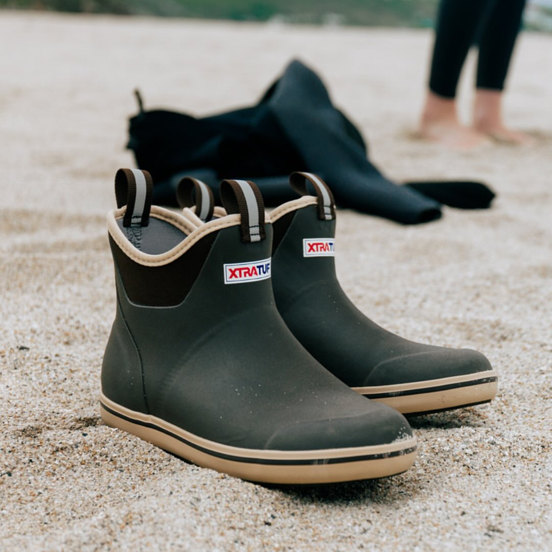 Pair of black rubber rain boots with beige soles and pull straps, placed on sandy ground at the beach.