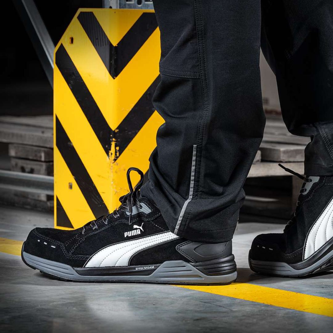 Close-up of a person's feet wearing black and white Puma sneakers, standing on a gray floor near a yellow and black caution sign in an industrial setting.